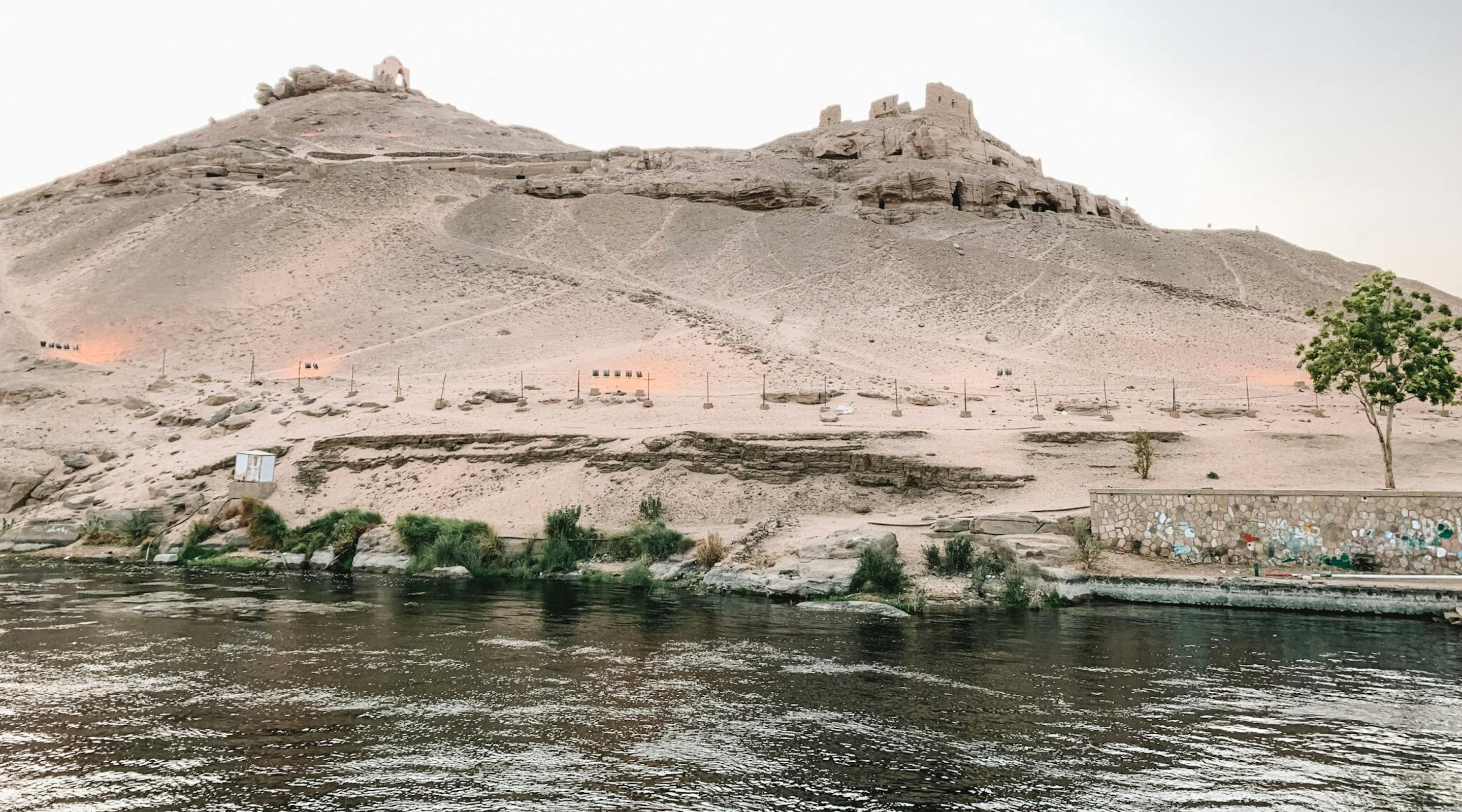 Low desert hills and small buildings sit across the Nile near Aswan, reflected in rippling water under pale light.