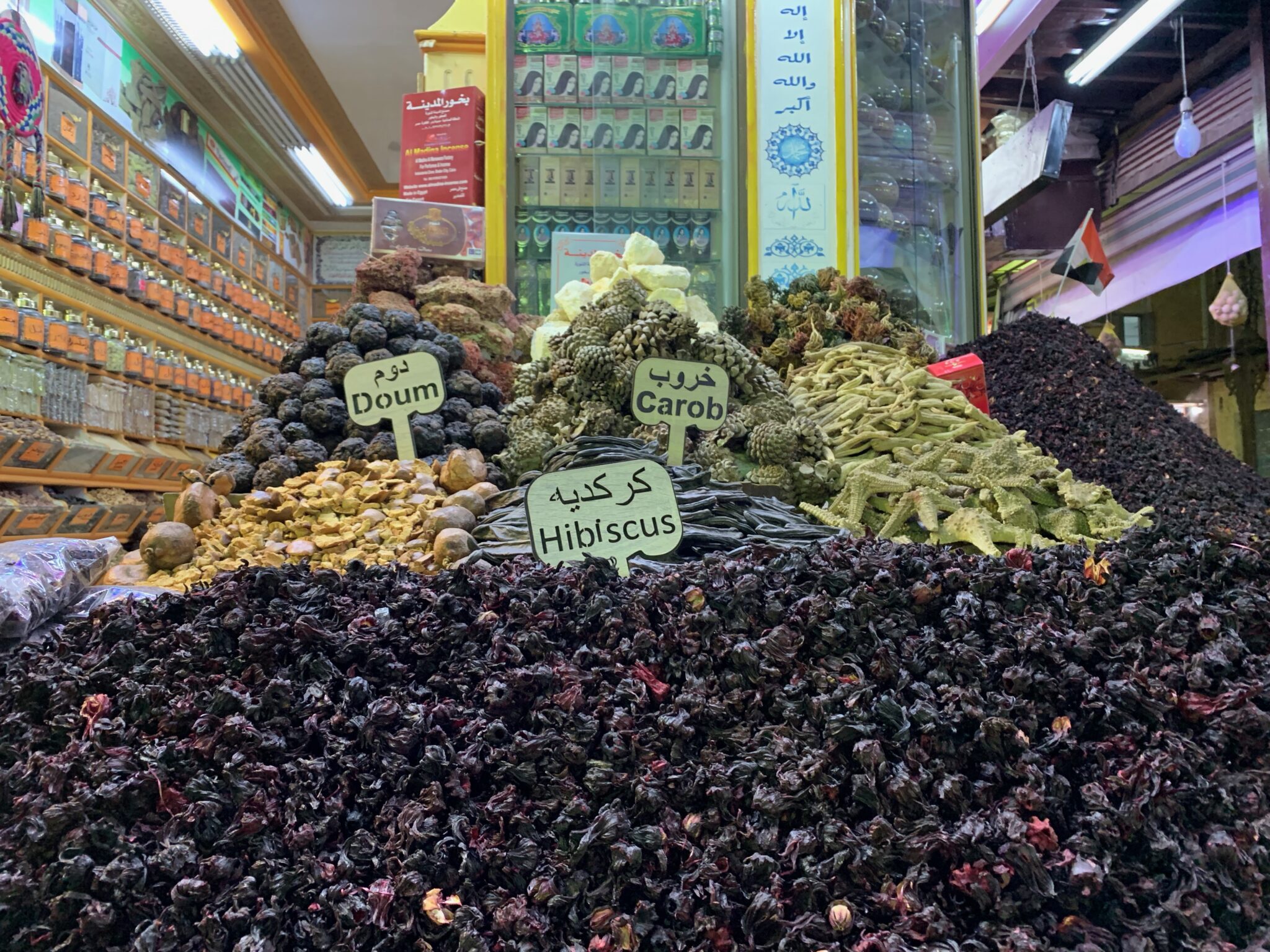 Piles of dried hibiscus and herbs fill a market stall, with handwritten price signs and colorful shelves in the background.