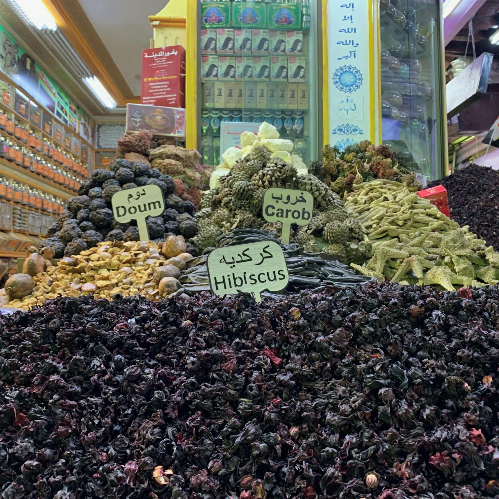 Piles of dried hibiscus and herbs fill a market stall, with handwritten price signs and colorful shelves in the background.