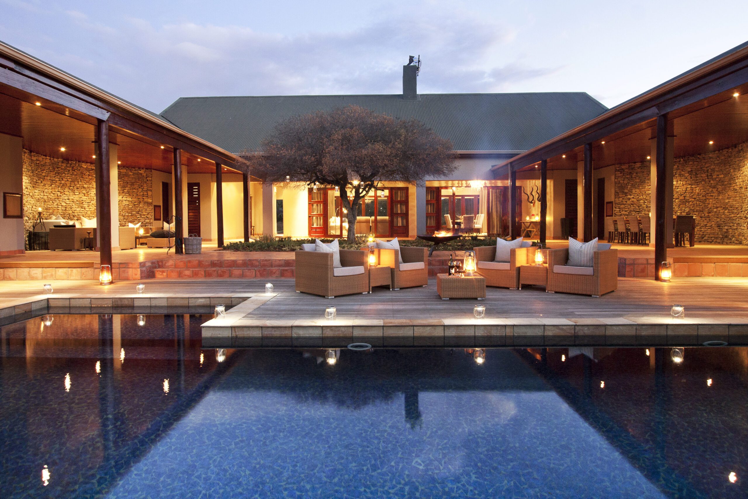 Lit courtyard pool reflects a modern lodge, with open-air lounges, stone walls, and warm lantern light at dusk.