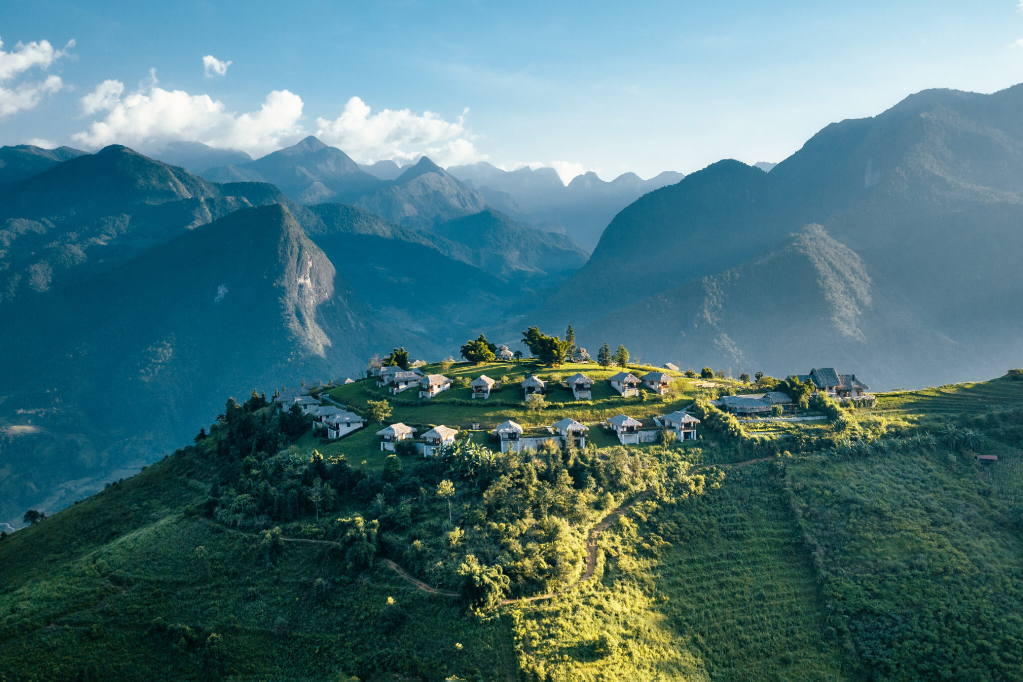 Mountain ridges frame a hilltop lodge complex with scattered villas and paths, lit by afternoon sun and drifting clouds.