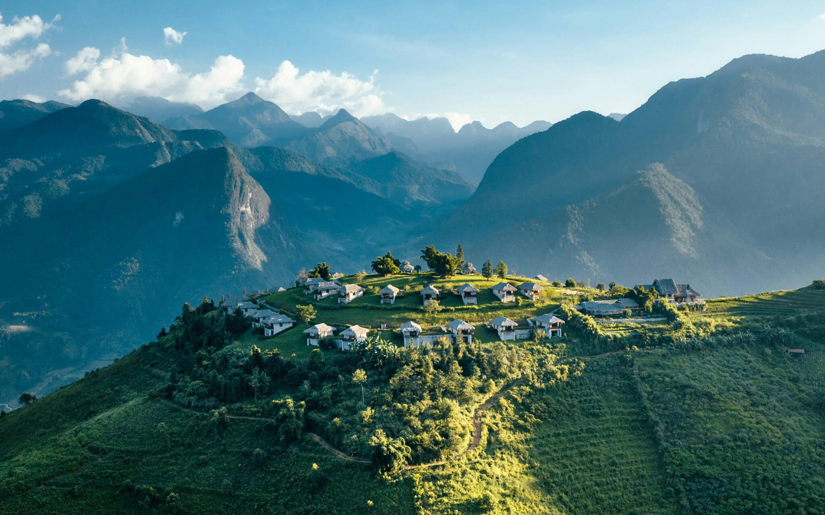 Mountain ridges frame a hilltop lodge complex with scattered villas and paths, lit by afternoon sun and drifting clouds.