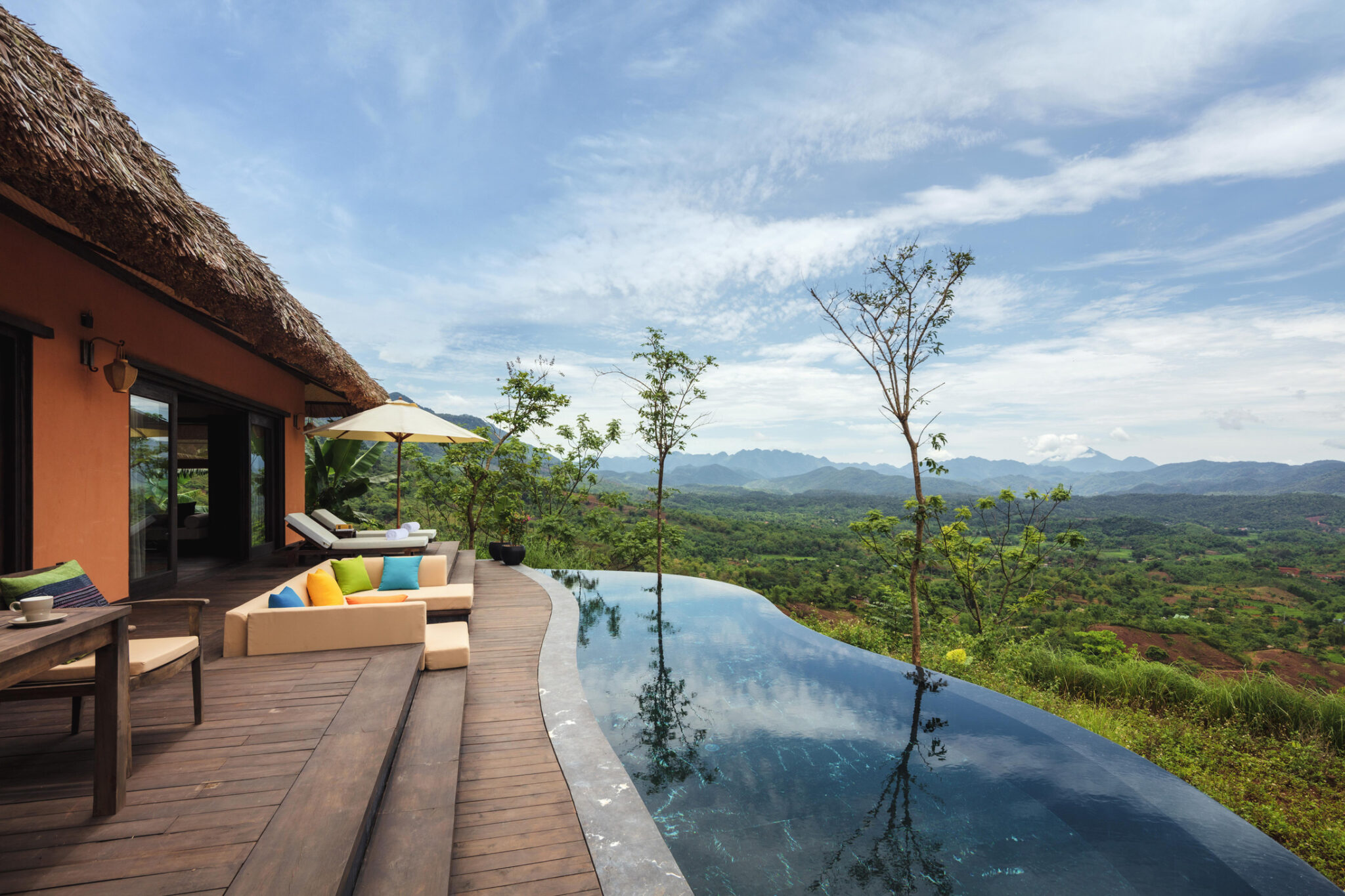 An infinity pool beside a villa terrace reflects gray clouds, looking out over forested hills and distant mountains.