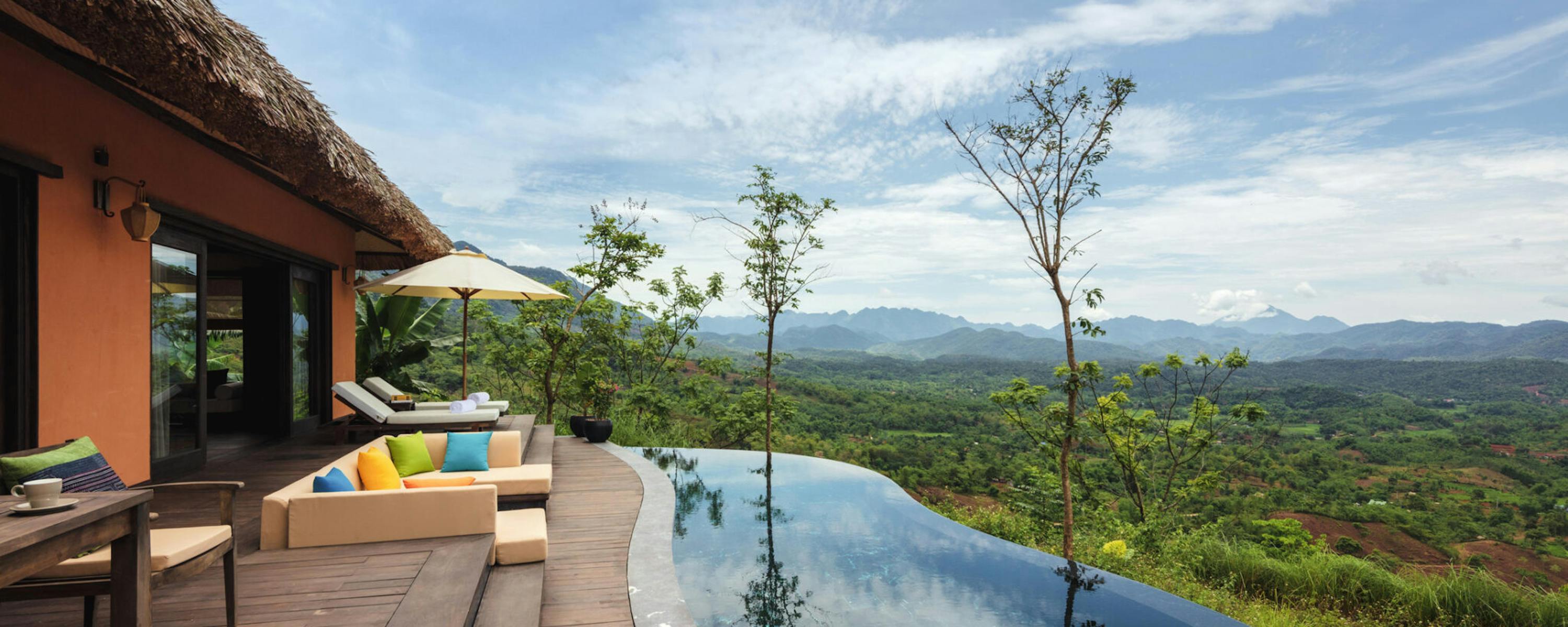 An infinity pool beside a villa terrace reflects gray clouds, looking out over forested hills and distant mountains.