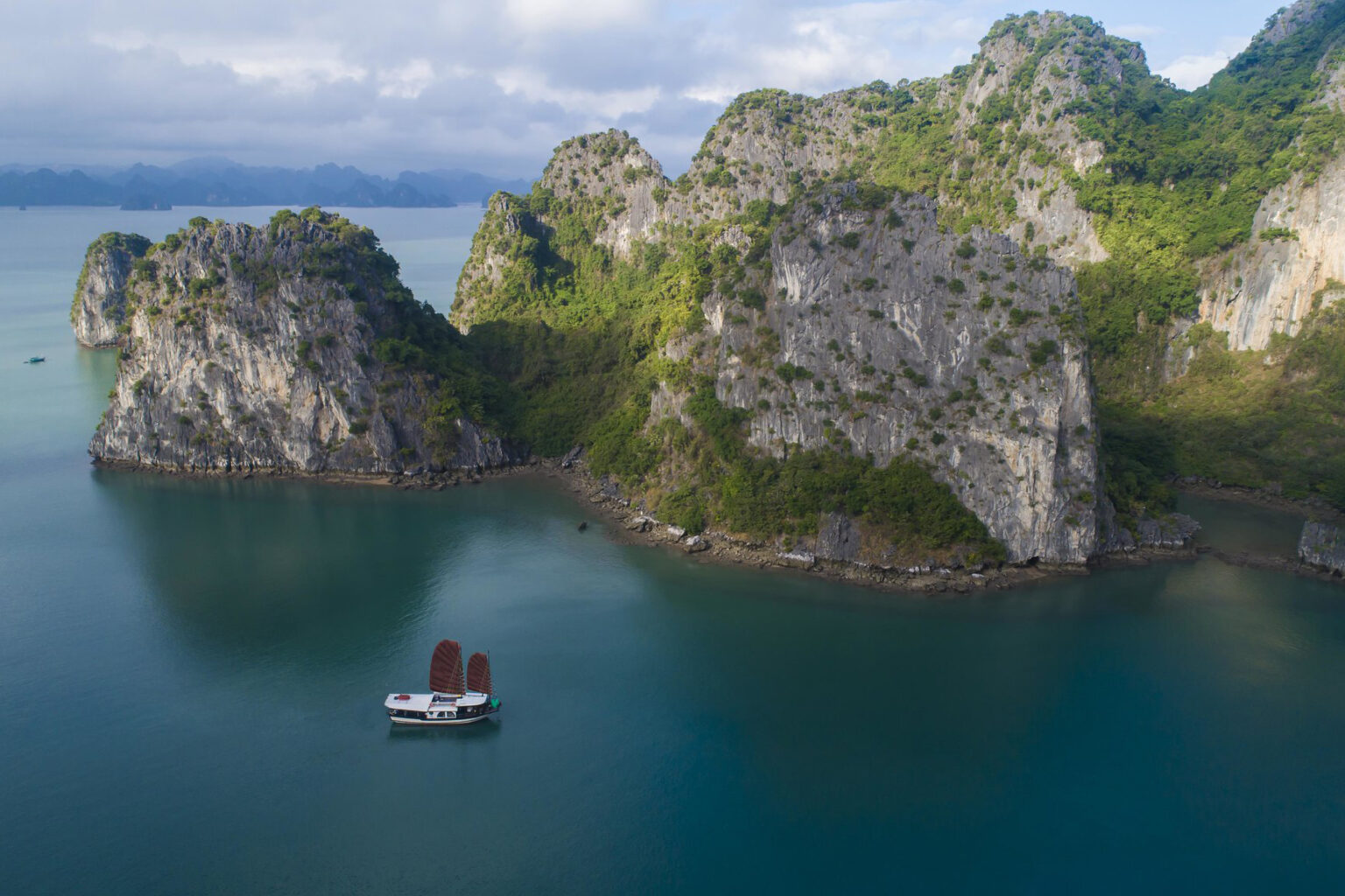 A small cruise boat sails through emerald water below steep limestone cliffs, with misty islands in the distance.