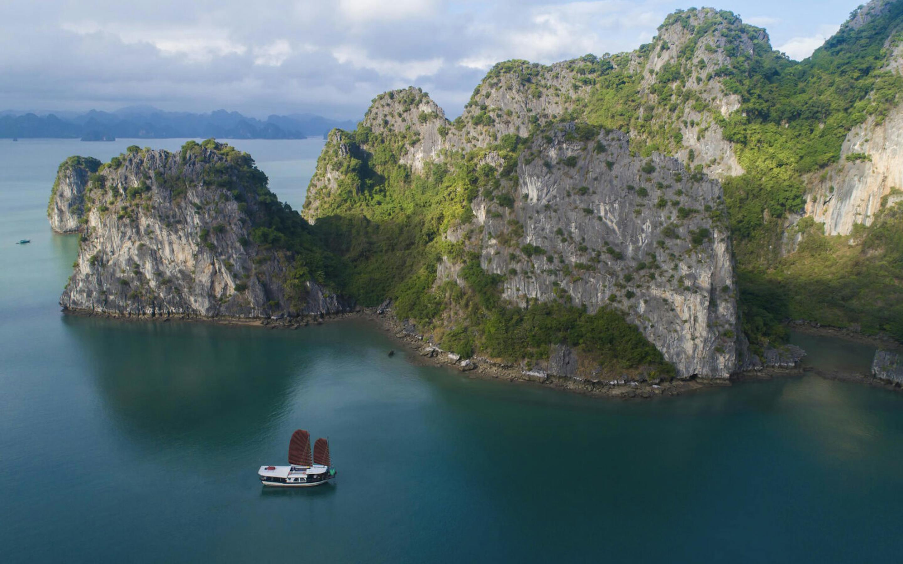 A small cruise boat sails through emerald water below steep limestone cliffs, with misty islands in the distance.