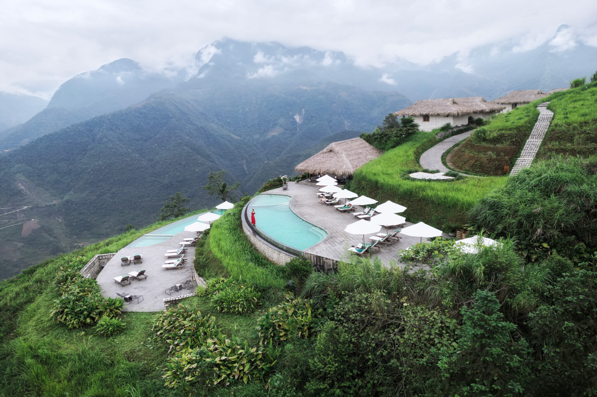 A curved infinity pool and terrace overlook misty mountains, with lounge chairs arranged along the ridge-side deck.