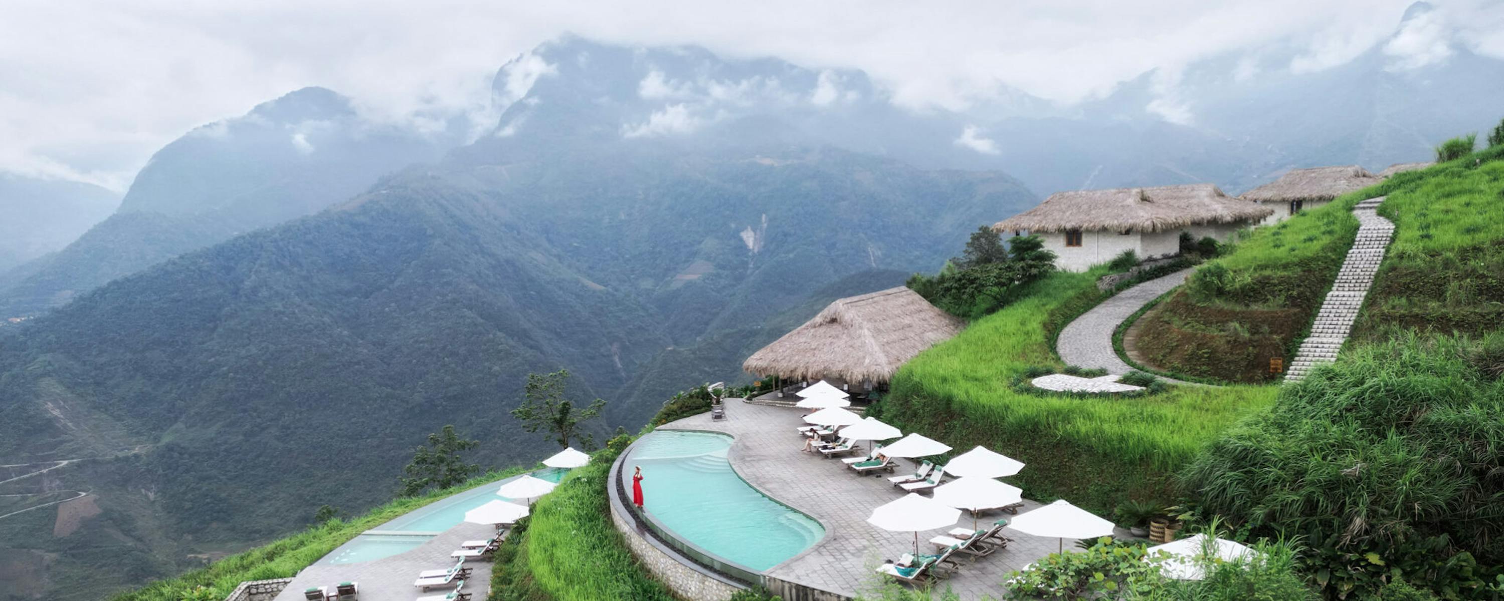 A curved infinity pool and terrace overlook misty mountains, with lounge chairs arranged along the ridge-side deck.