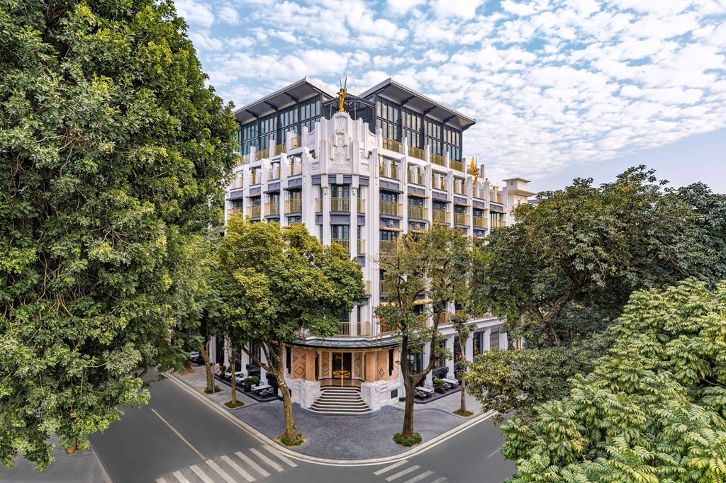 A modern hotel facade rises above a street corner, framed by leafy trees beneath a sky of scattered white clouds.