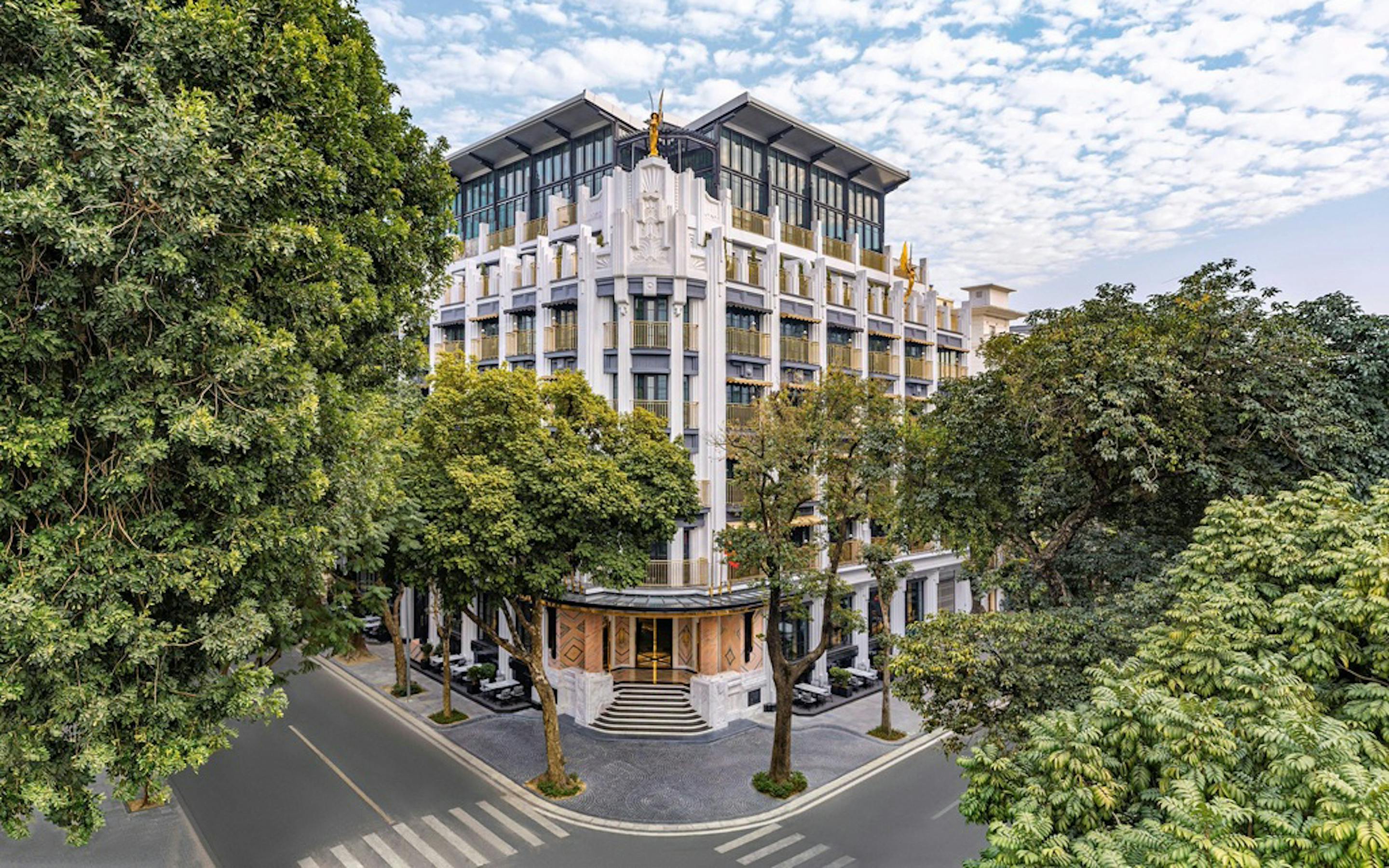 A modern hotel facade rises above a street corner, framed by leafy trees beneath a sky of scattered white clouds.