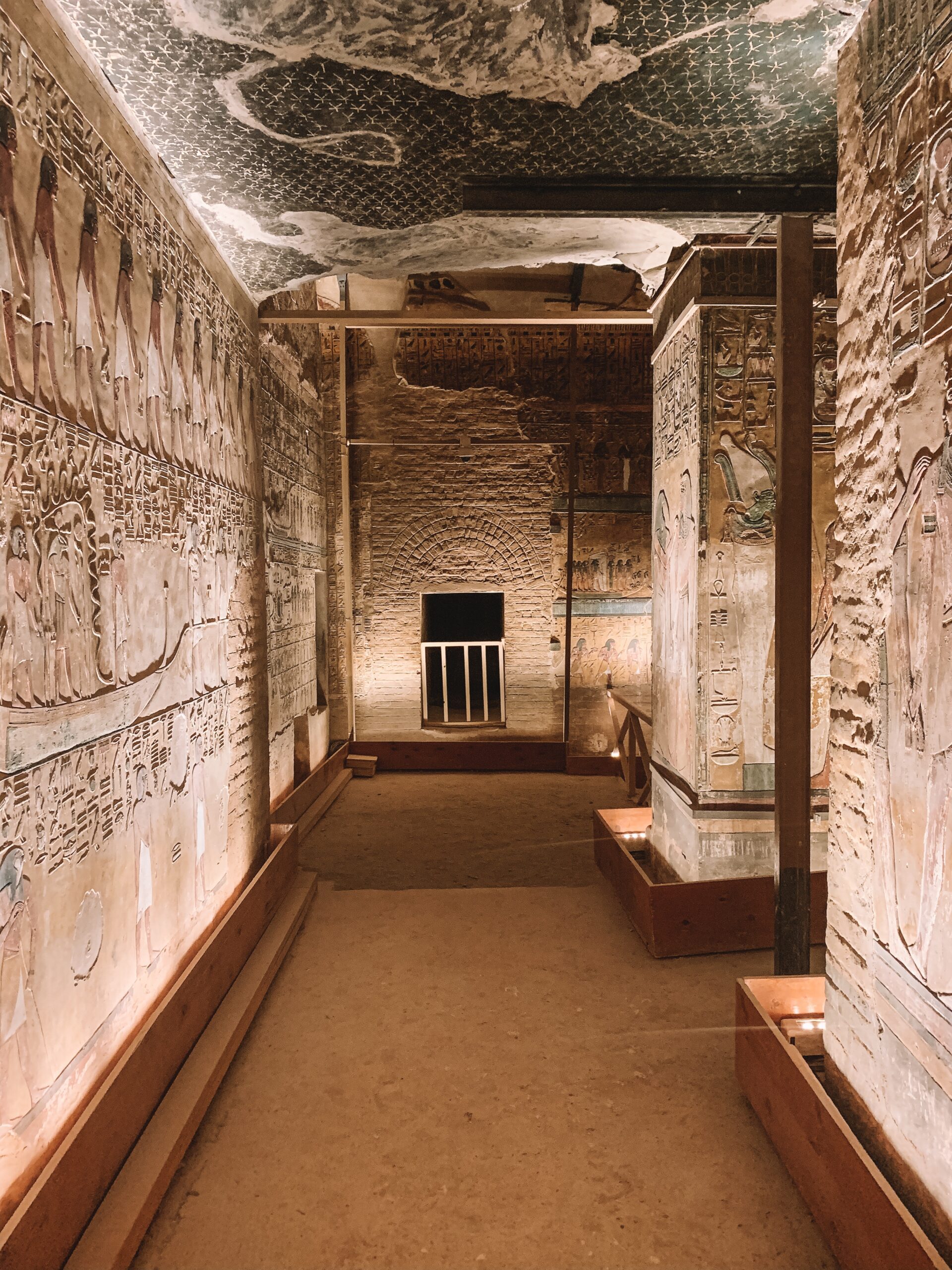 Lit, narrow tomb corridor with hieroglyph-lined walls leads to a doorway, with a carved ceiling and stone floor.