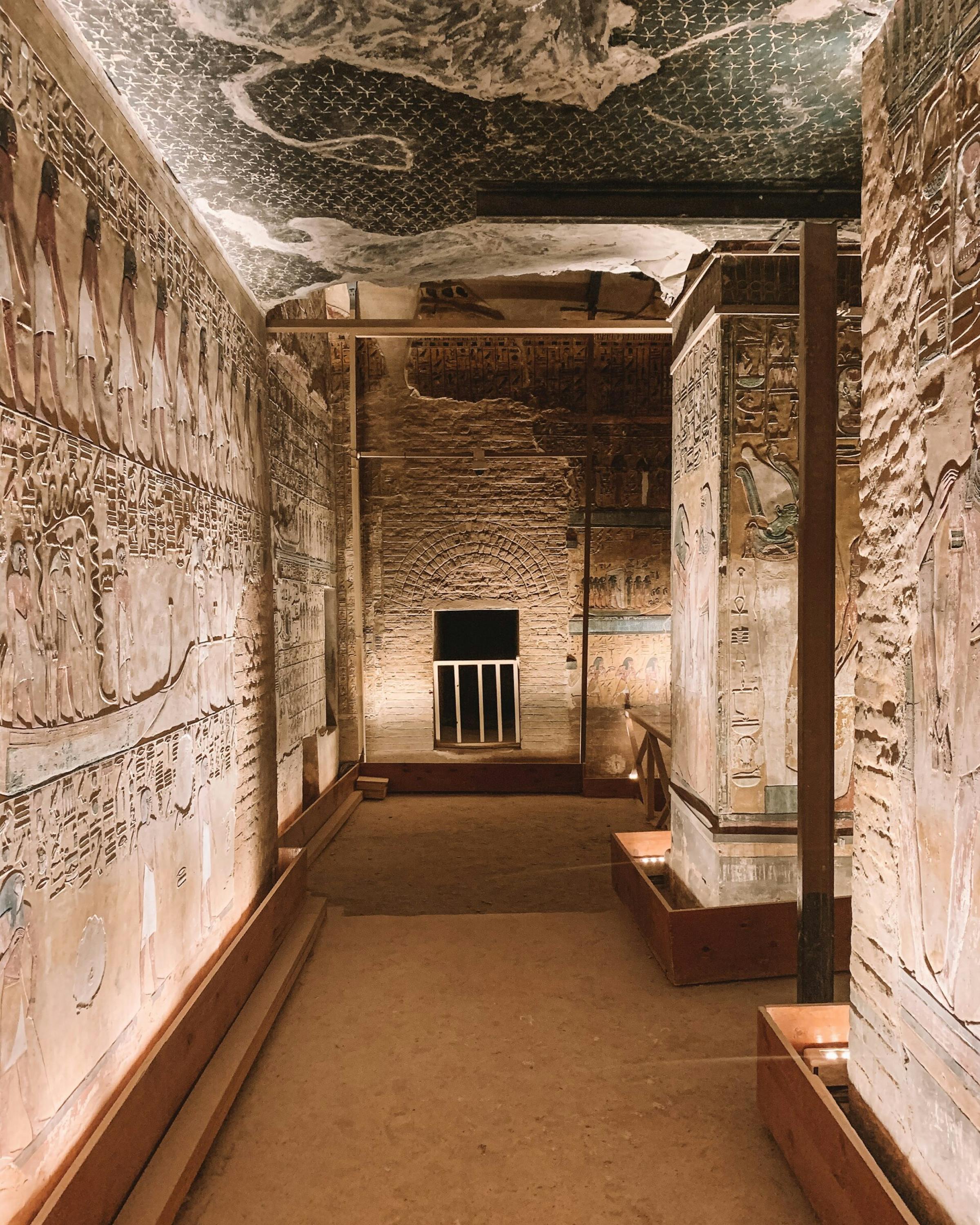 Lit, narrow tomb corridor with hieroglyph-lined walls leads to a doorway, with a carved ceiling and stone floor.