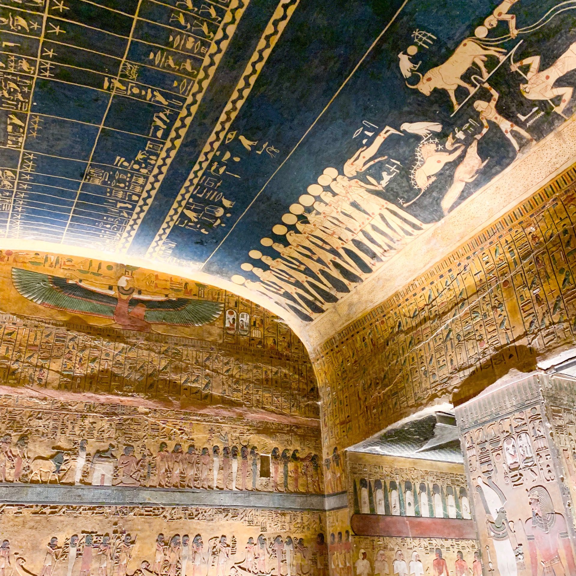Ornate tomb ceiling painted with stars and figures arches over murals, glowing in golden light in the Valley of Kings.