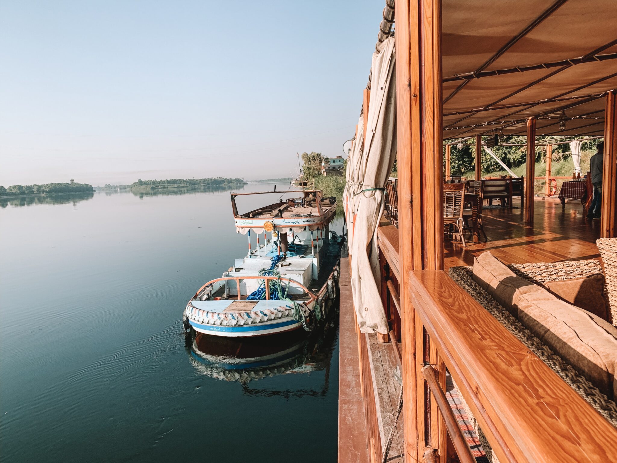 A river cruise boat moors beside a wooden deck, reflected in quiet still water as other boats line the bank.