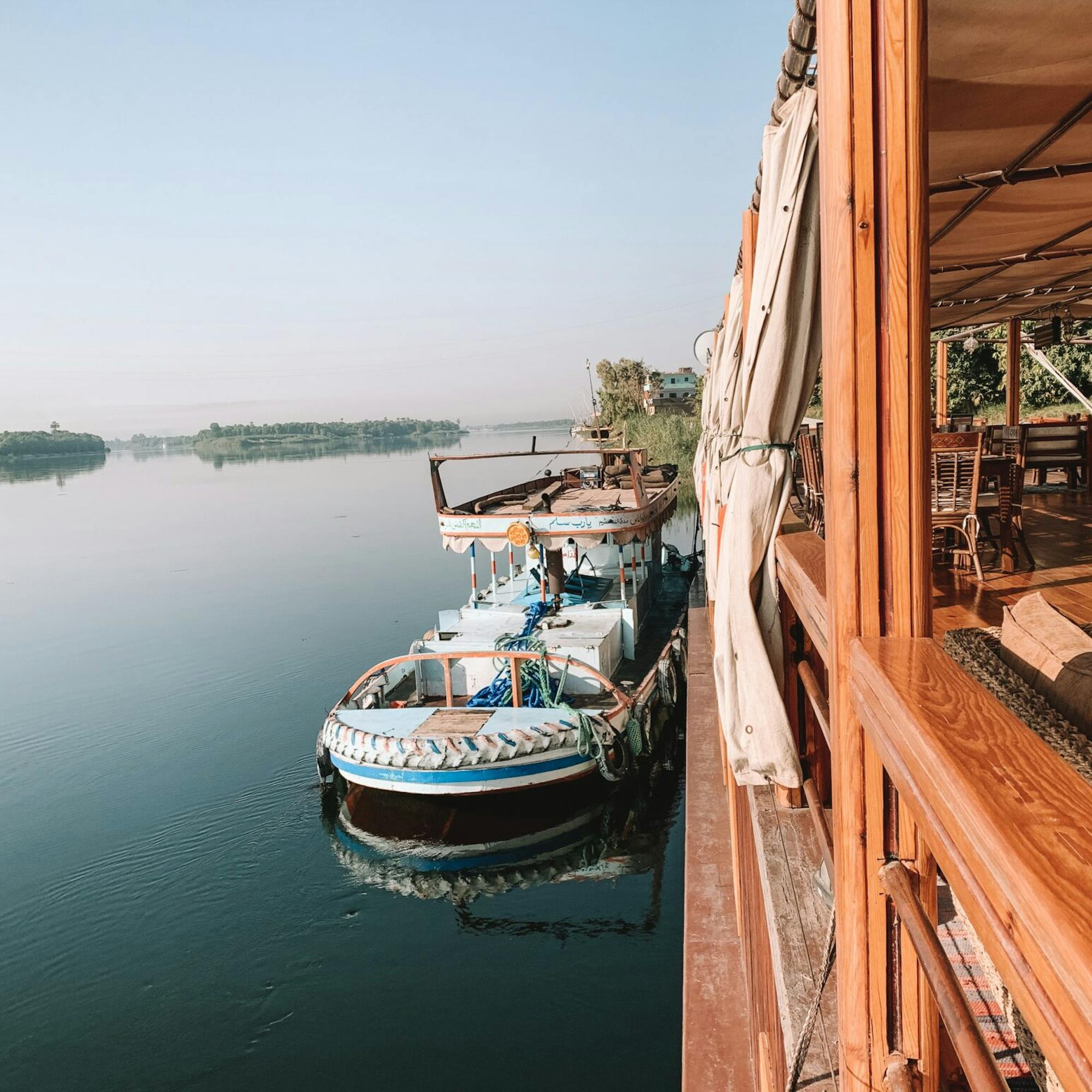 A river cruise boat moors beside a wooden deck, reflected in quiet still water as other boats line the bank.