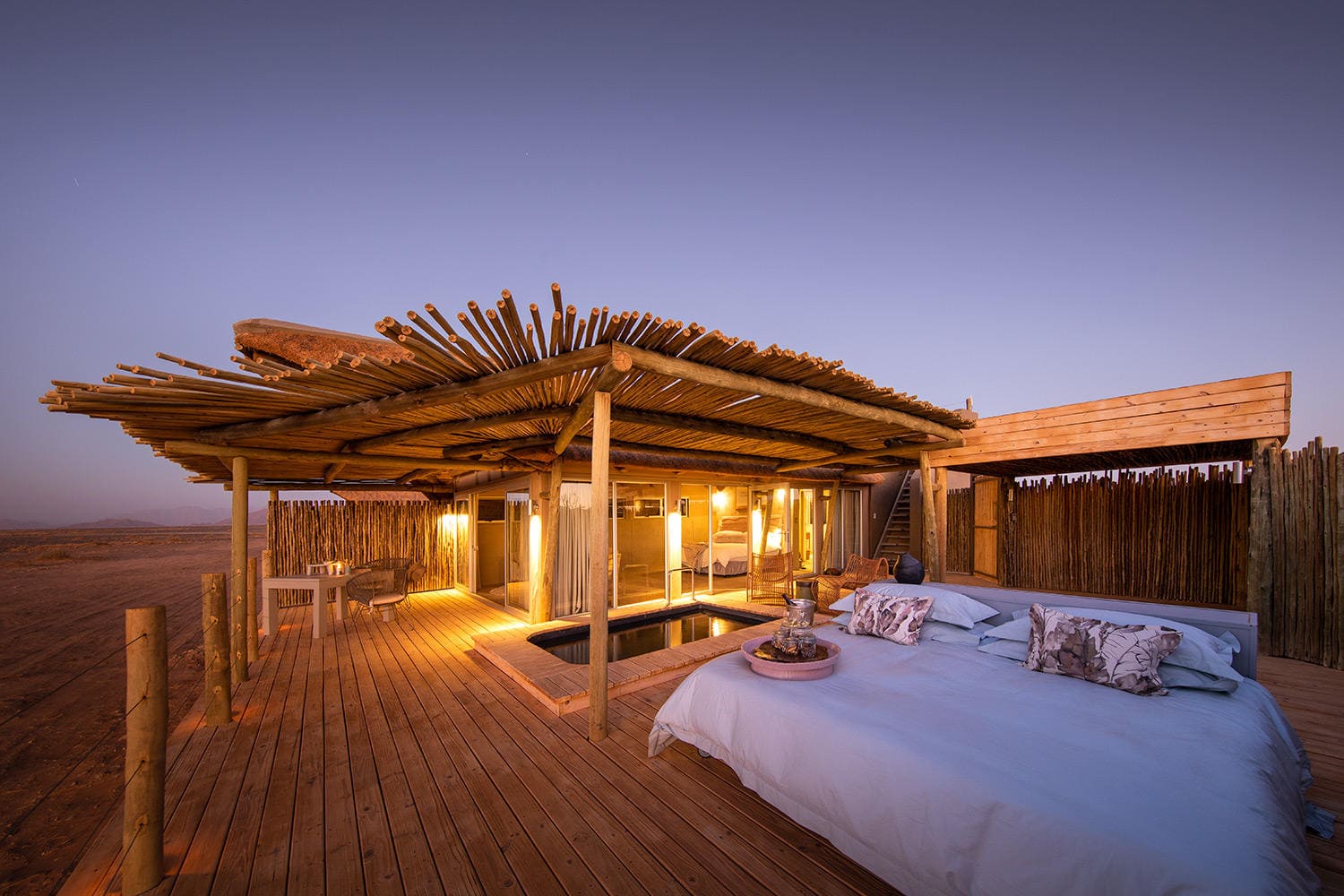 An open-air bed glows on a rooftop deck at dusk, with desert dunes nearby and soft lantern light around it.