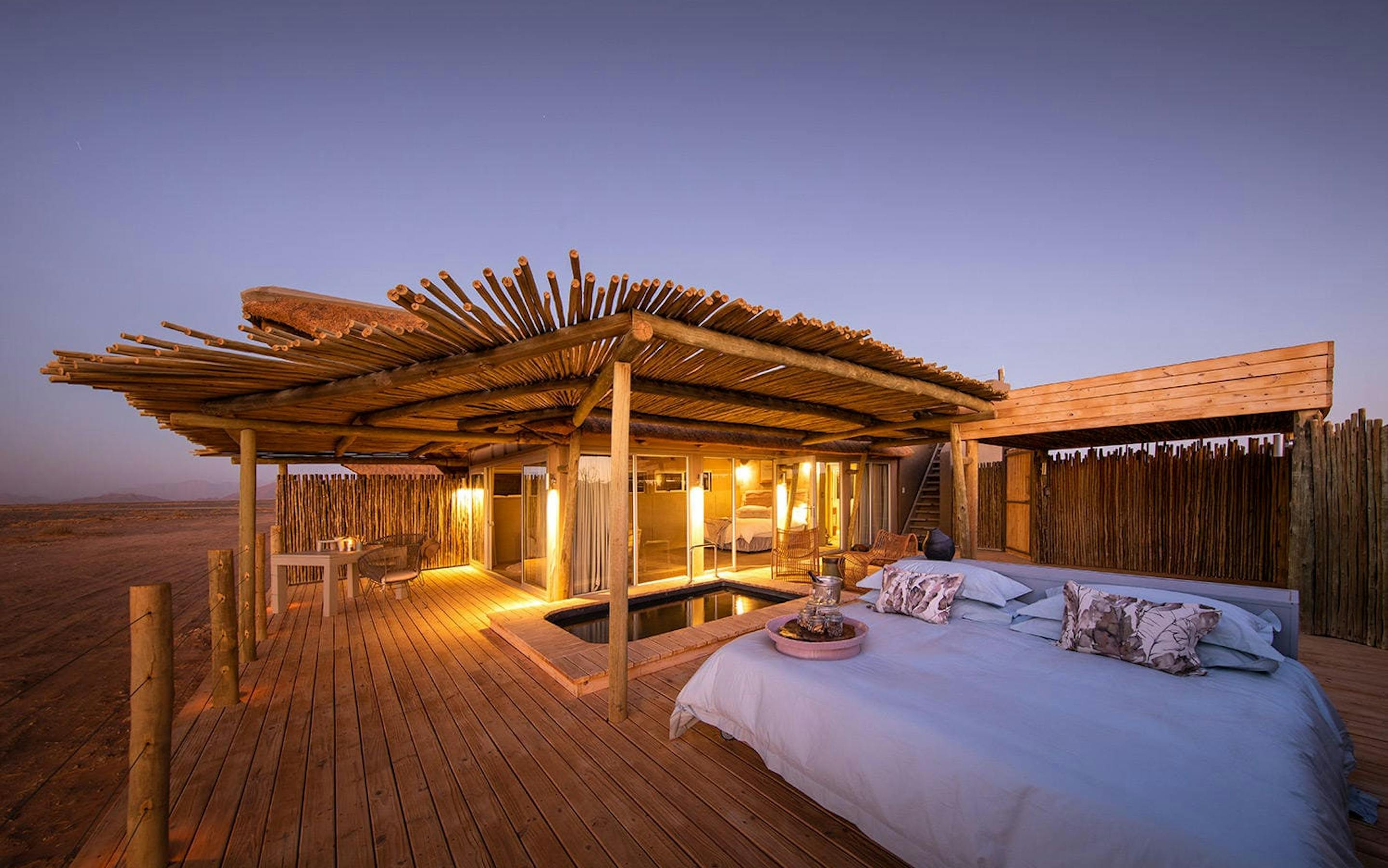 An open-air bed glows on a rooftop deck at dusk, with desert dunes nearby and soft lantern light around it.