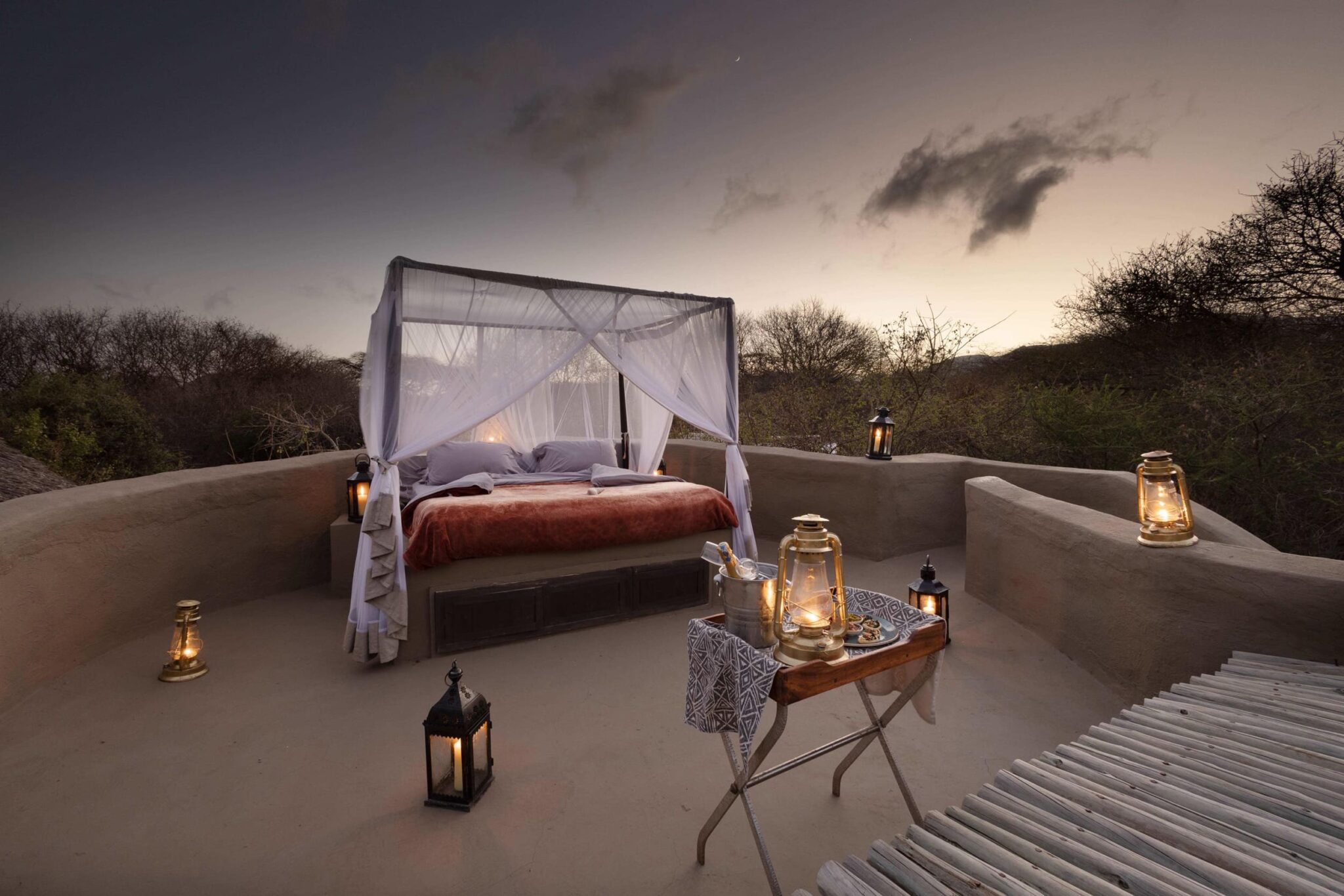 An outdoor canopy bed is set on a terrace with lanterns, facing open plains and a dramatic dusk sky in the distance.