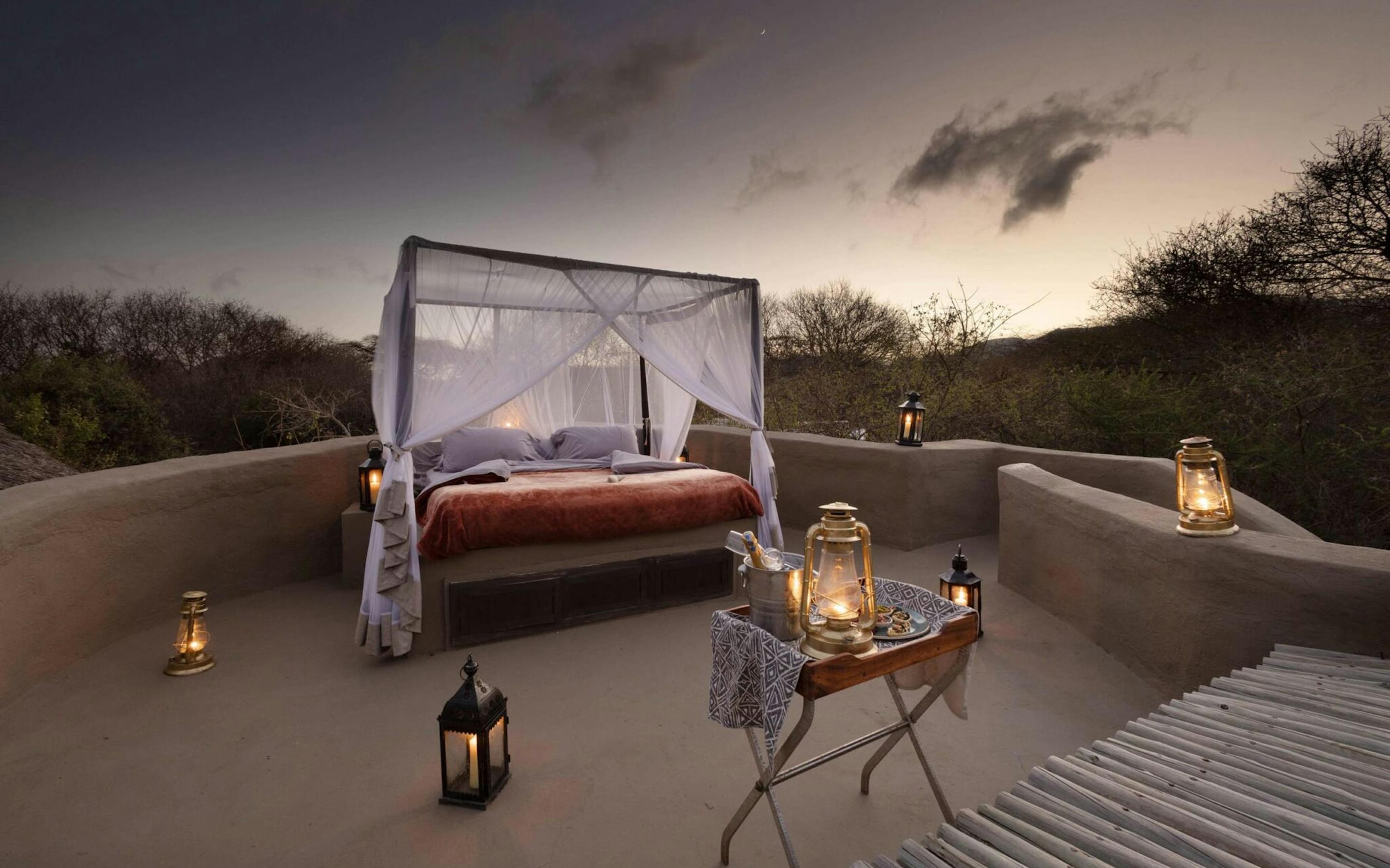 An outdoor canopy bed is set on a terrace with lanterns, facing open plains and a dramatic dusk sky in the distance.