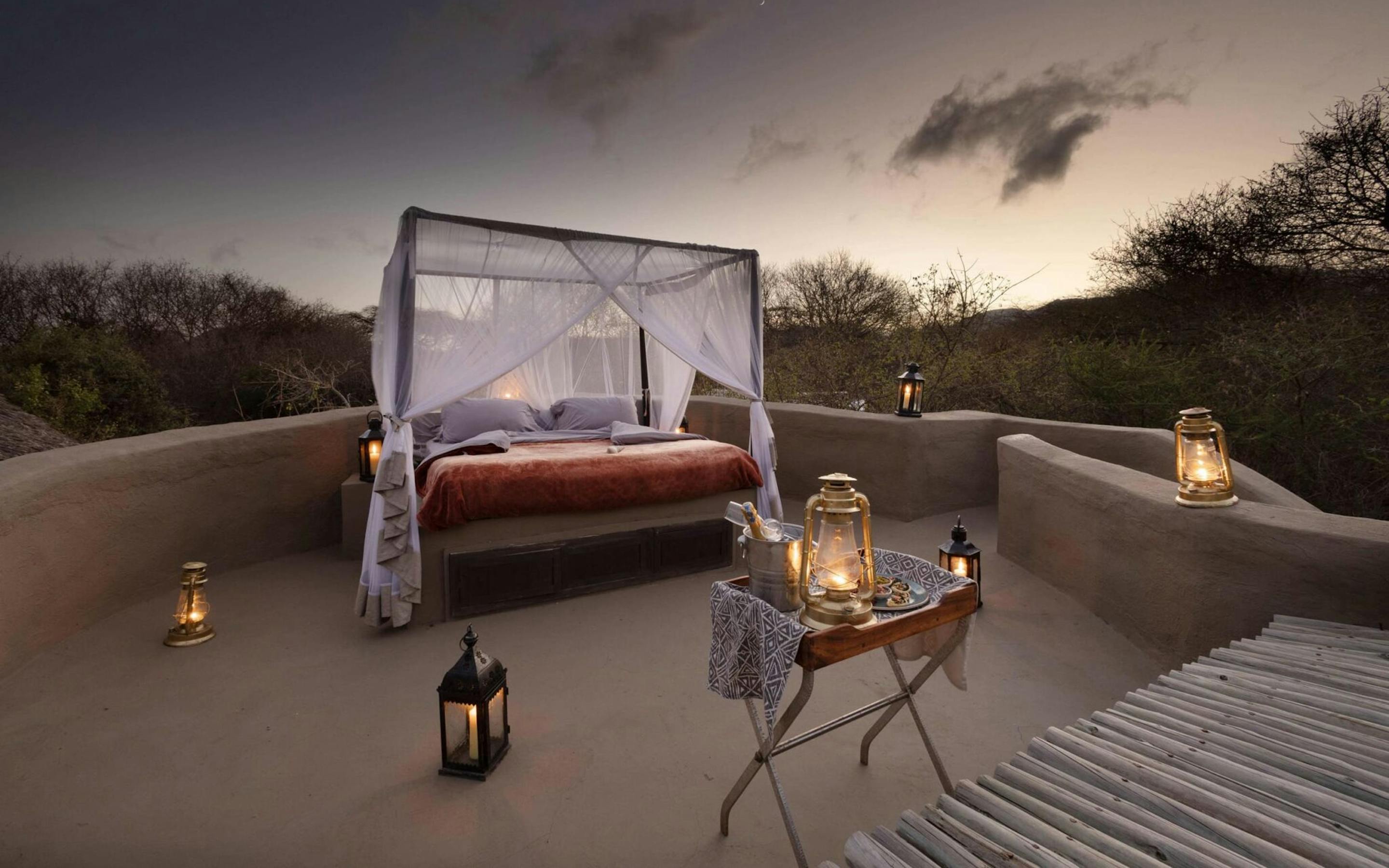 An outdoor canopy bed is set on a terrace with lanterns, facing open plains and a dramatic dusk sky in the distance.