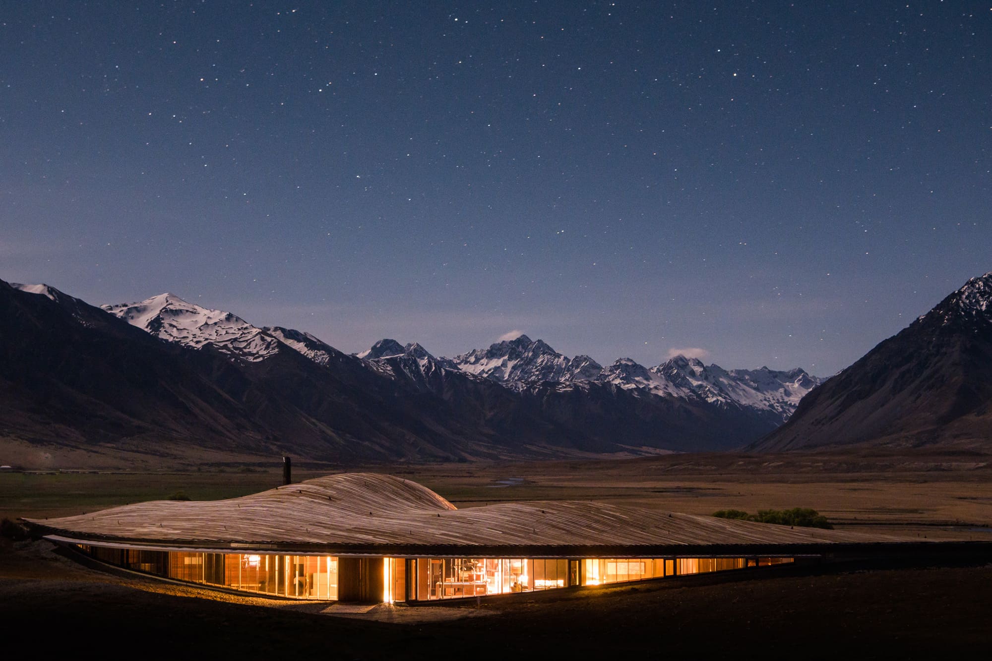 A lodge glows warmly in a mountain valley at night, with snowcapped peaks silhouetted under a clear dark sky.
