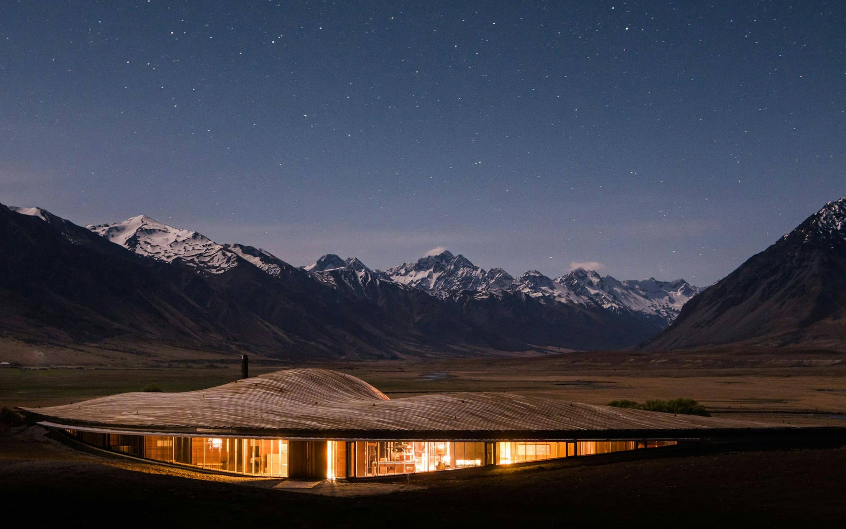A lodge glows warmly in a mountain valley at night, with snowcapped peaks silhouetted under a clear dark sky.