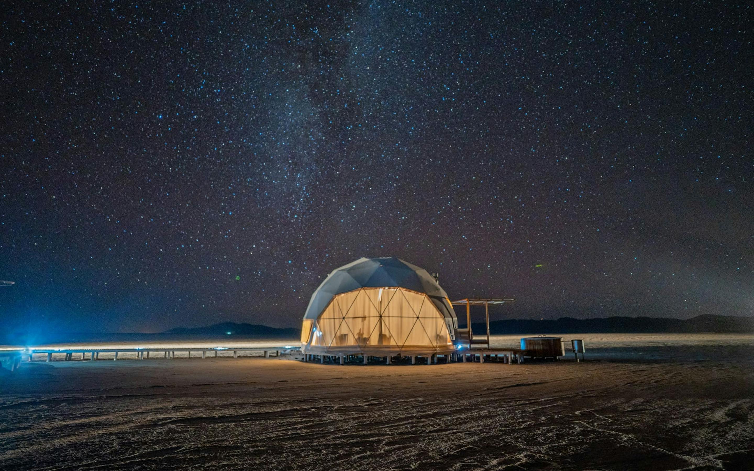 A lit geodesic dome sits on the Salinas Grandes salt flat as the Milky Way stretches across a starry sky.
