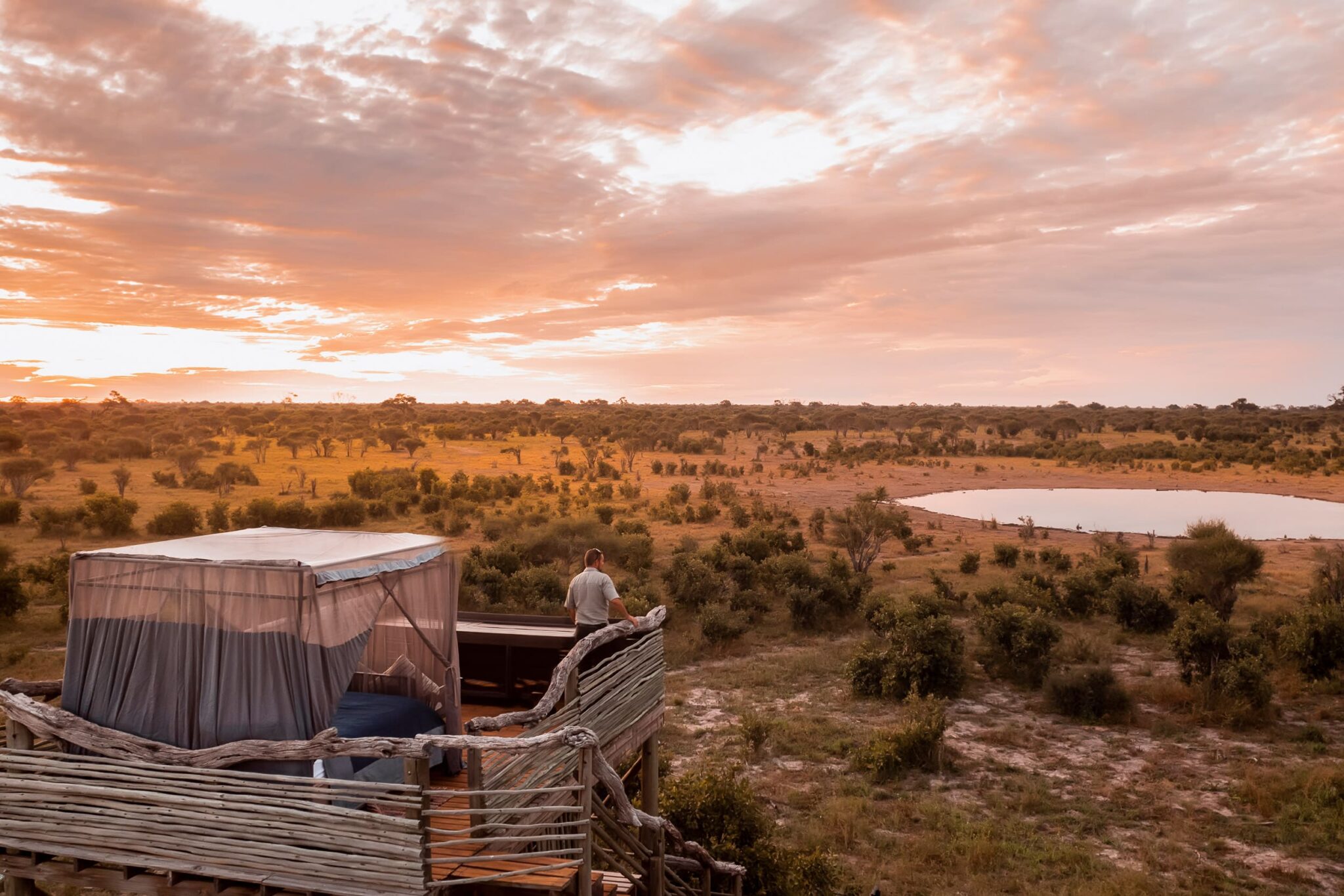 A canvas tent and deck overlook Okavango floodplains at sunset, with orange light reflecting on winding water channels.