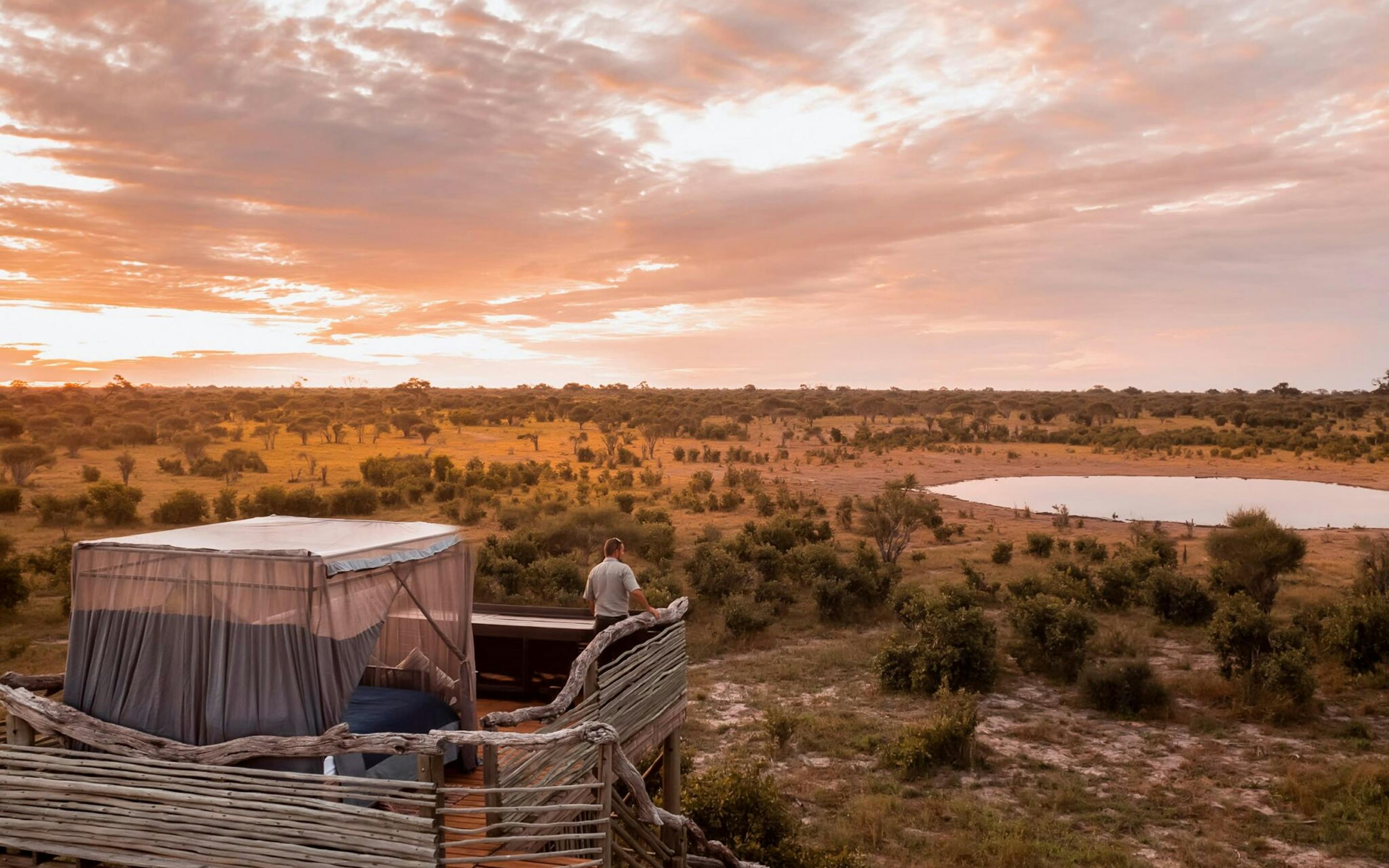 A canvas tent and deck overlook Okavango floodplains at sunset, with orange light reflecting on winding water channels.