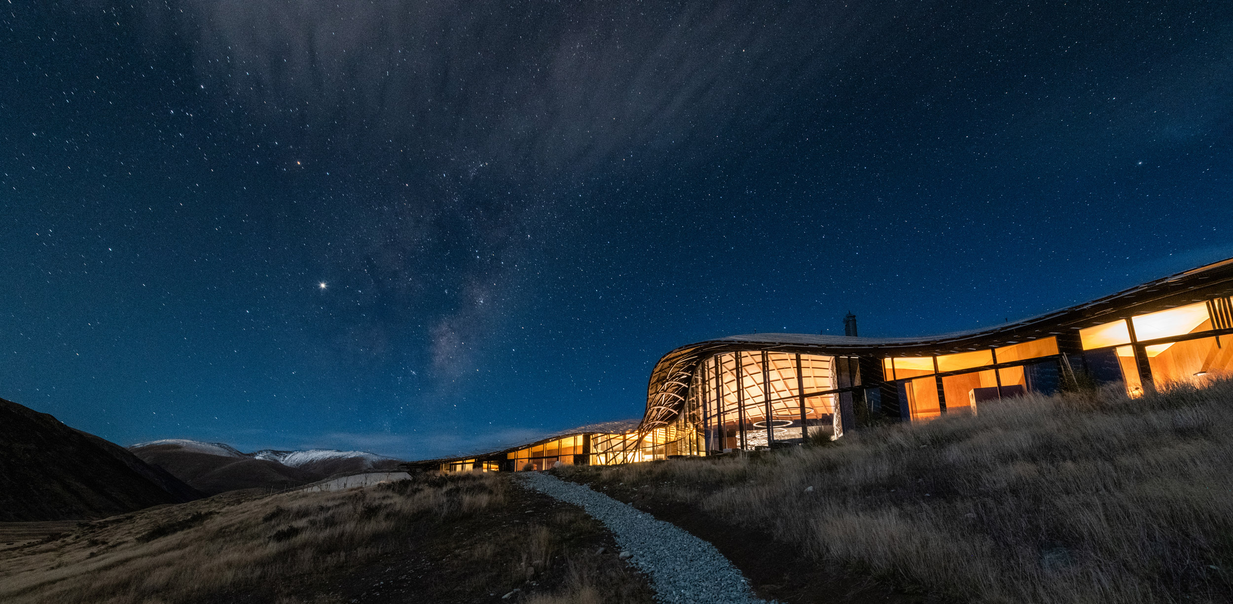 Modern lodge glows at night beneath a star-filled sky, with dark hills and valley silhouettes in the distance.