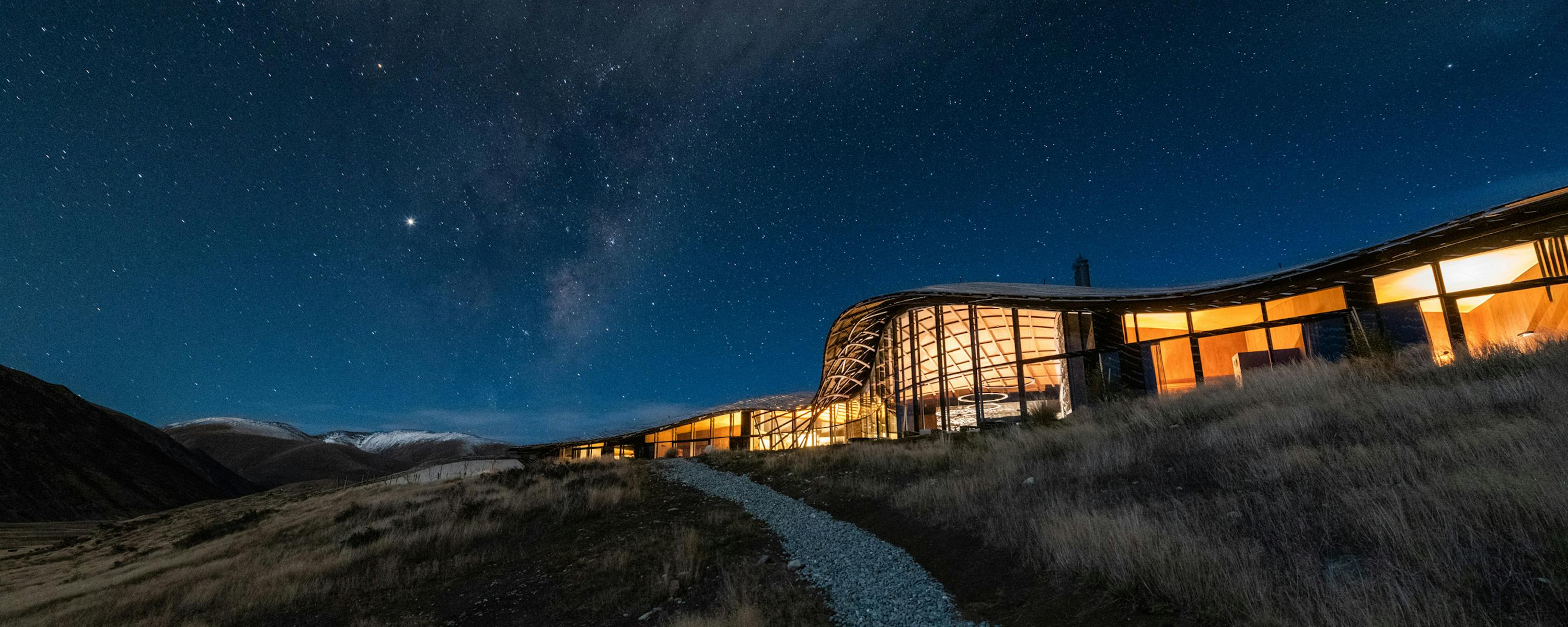 Modern lodge glows at night beneath a star-filled sky, with dark hills and valley silhouettes in the distance.