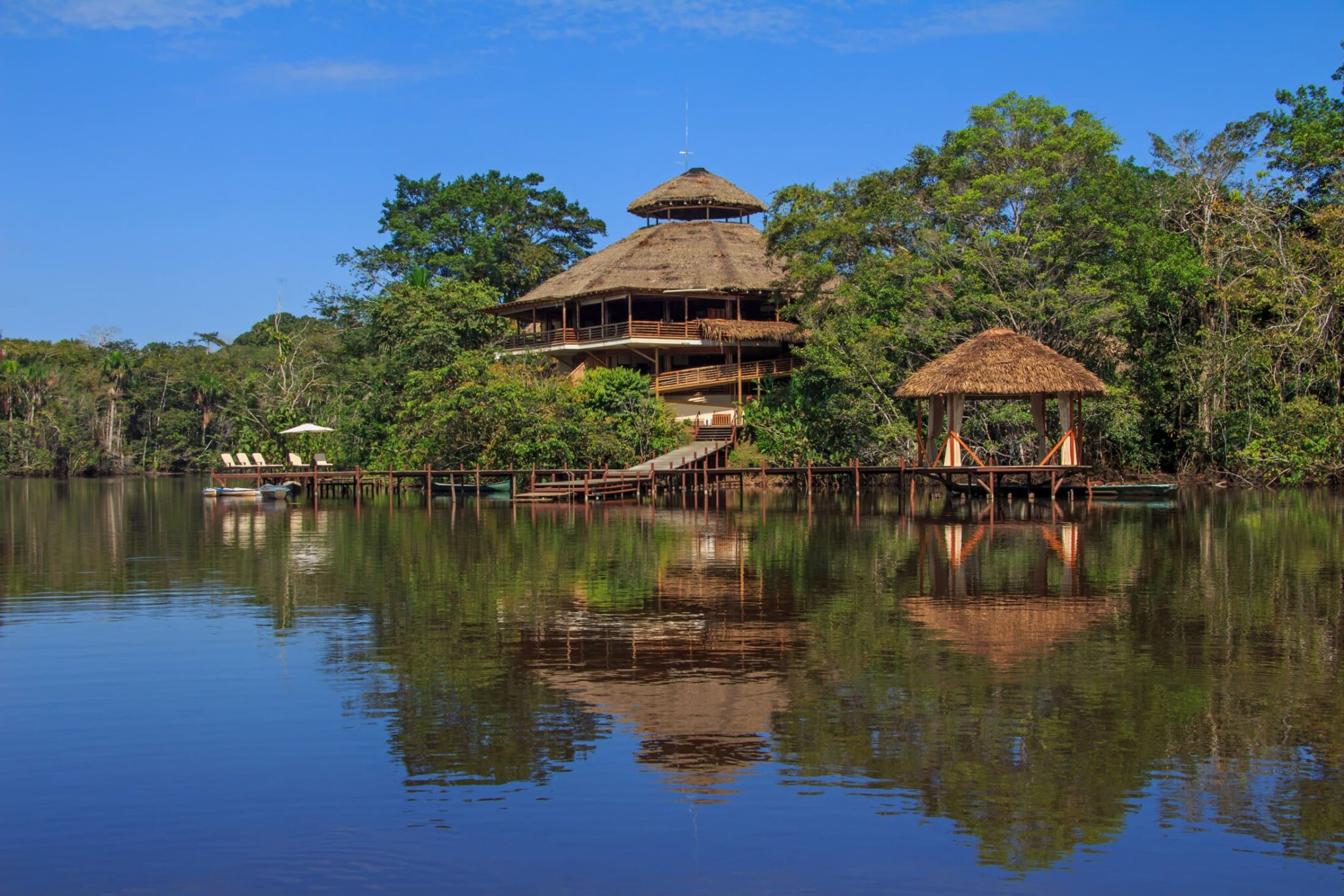 Thatched lodge buildings sit beside a calm lagoon in the Amazon, with lush rainforest reflected in the water.