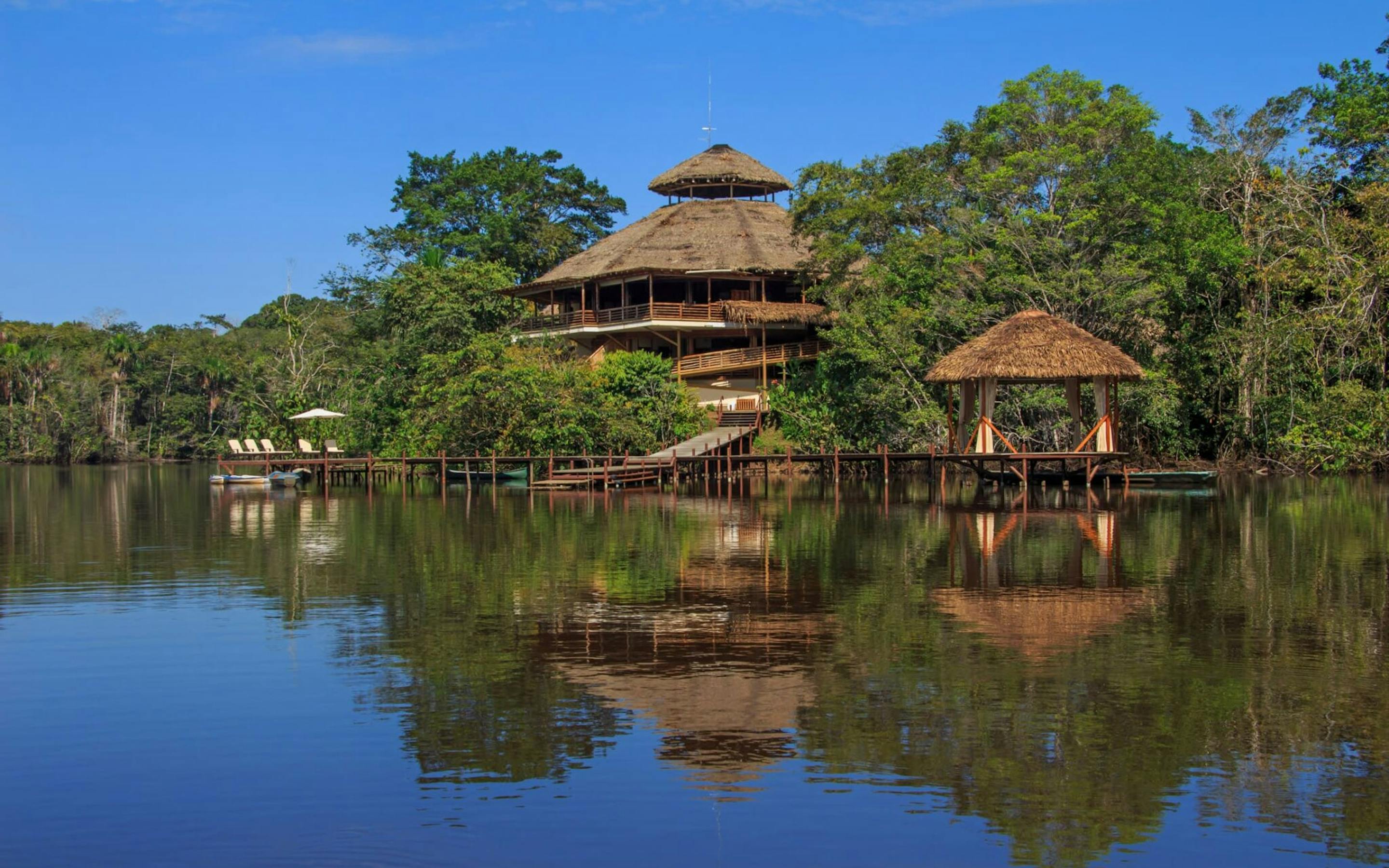 Thatched lodge buildings sit beside a calm lagoon in the Amazon, with lush rainforest reflected in the water.