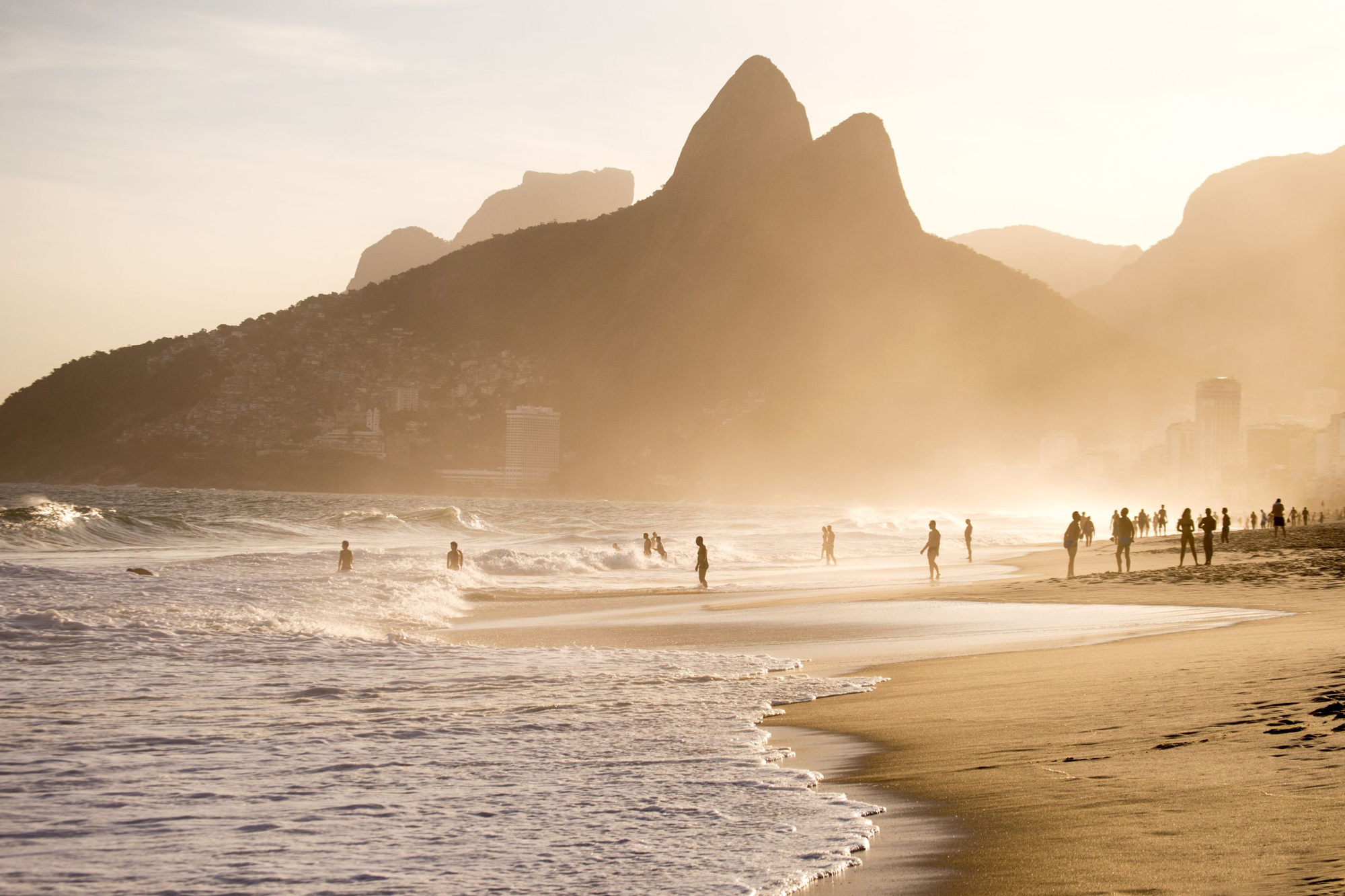 People stroll along Ipanema Beach at sunset, with soft waves, distant mountains, and a glowing golden sky beyond.