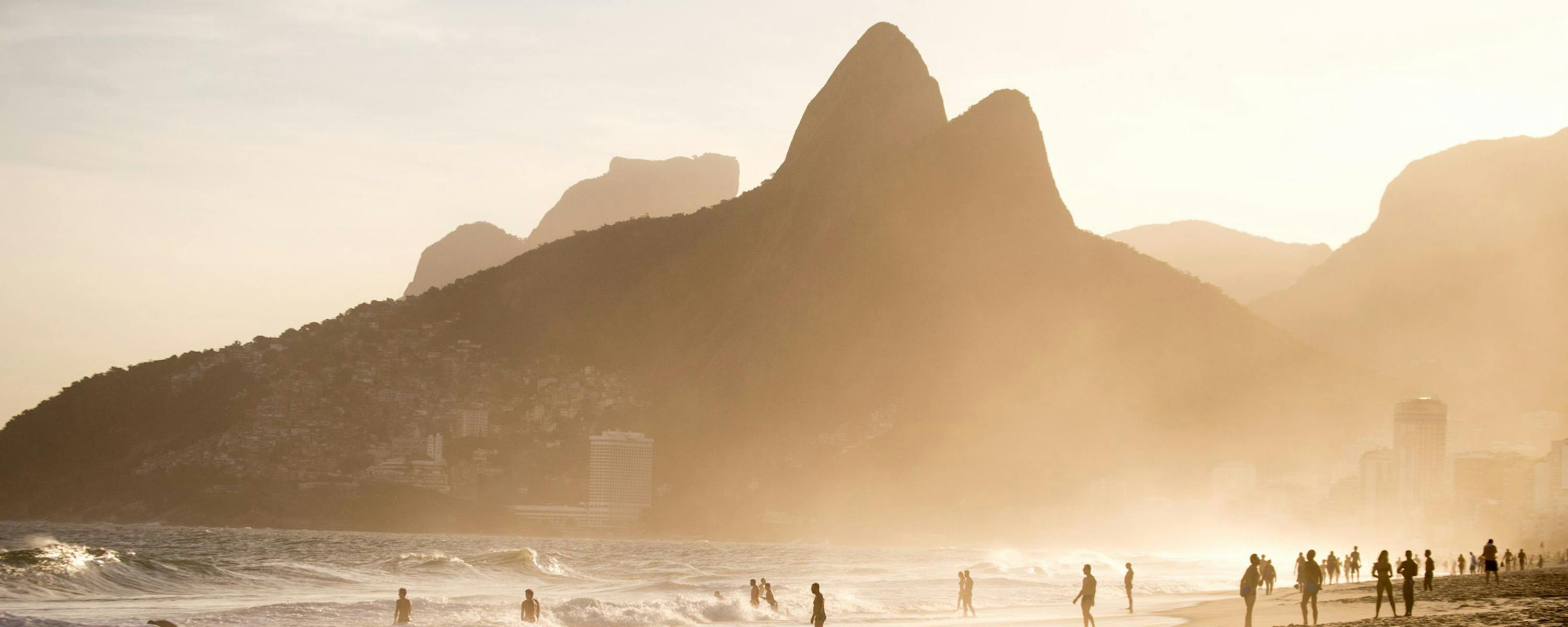 People stroll along Ipanema Beach at sunset, with soft waves, distant mountains, and a glowing golden sky beyond.