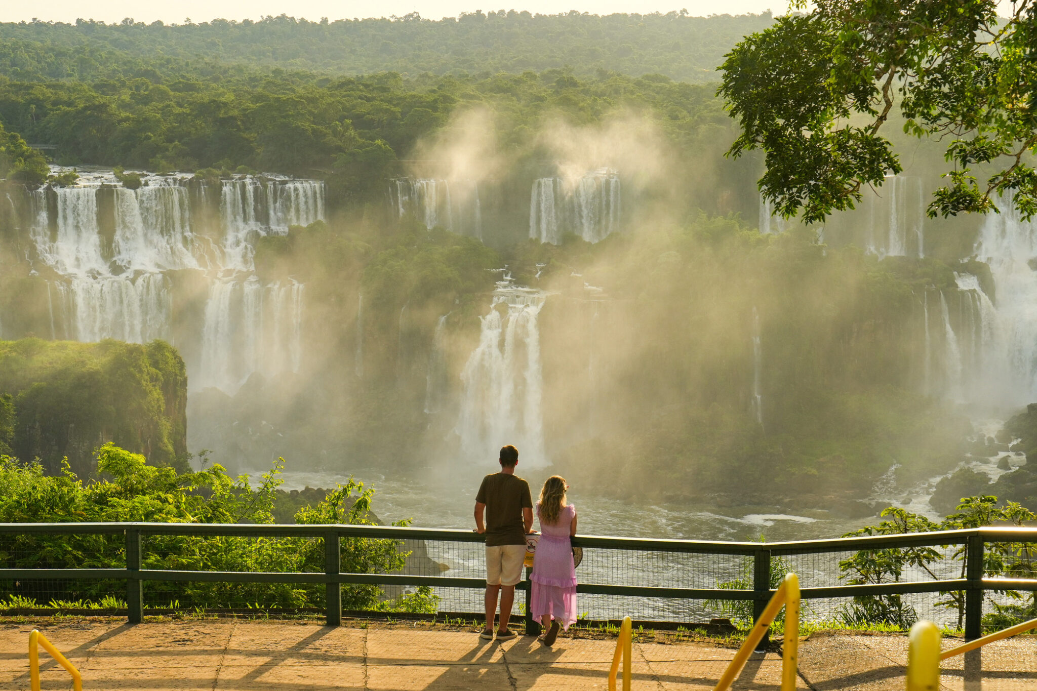 Two visitors stand at a railing overlooking Iguacu Falls, as mist rises from broad cascades in the gorge below.