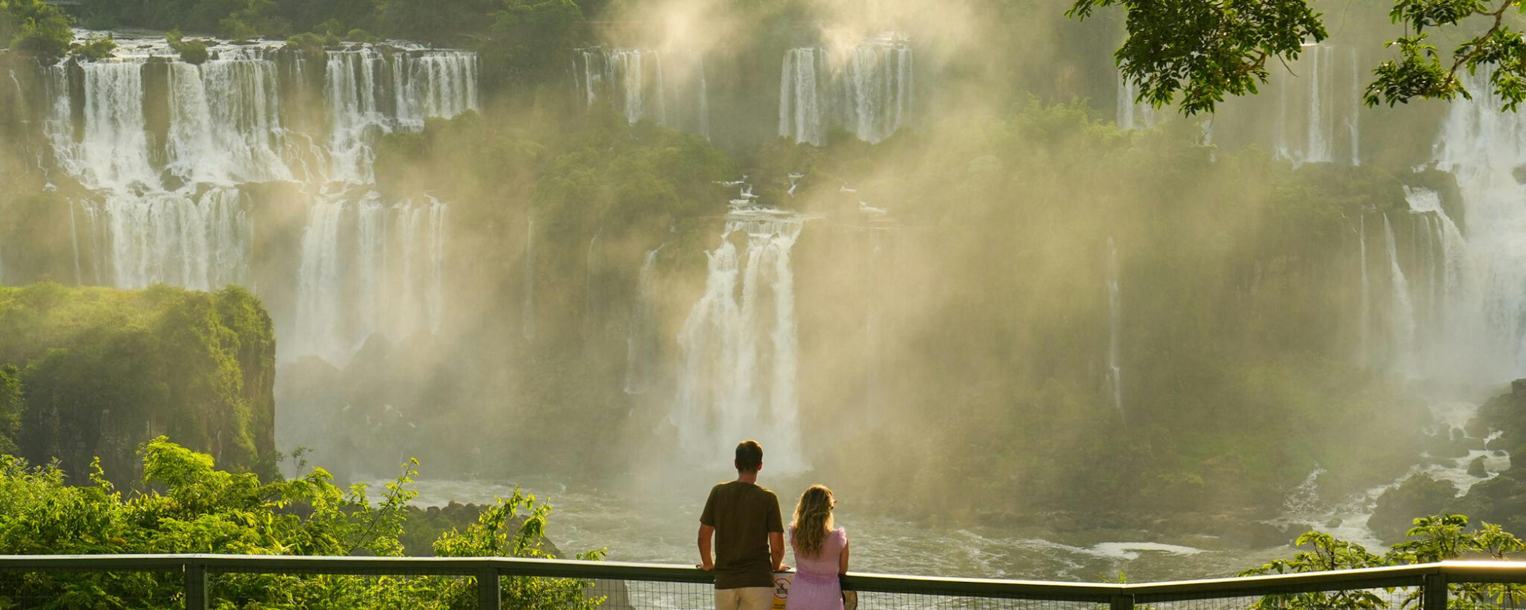 Two visitors stand at a railing overlooking Iguacu Falls, as mist rises from broad cascades in the gorge below.
