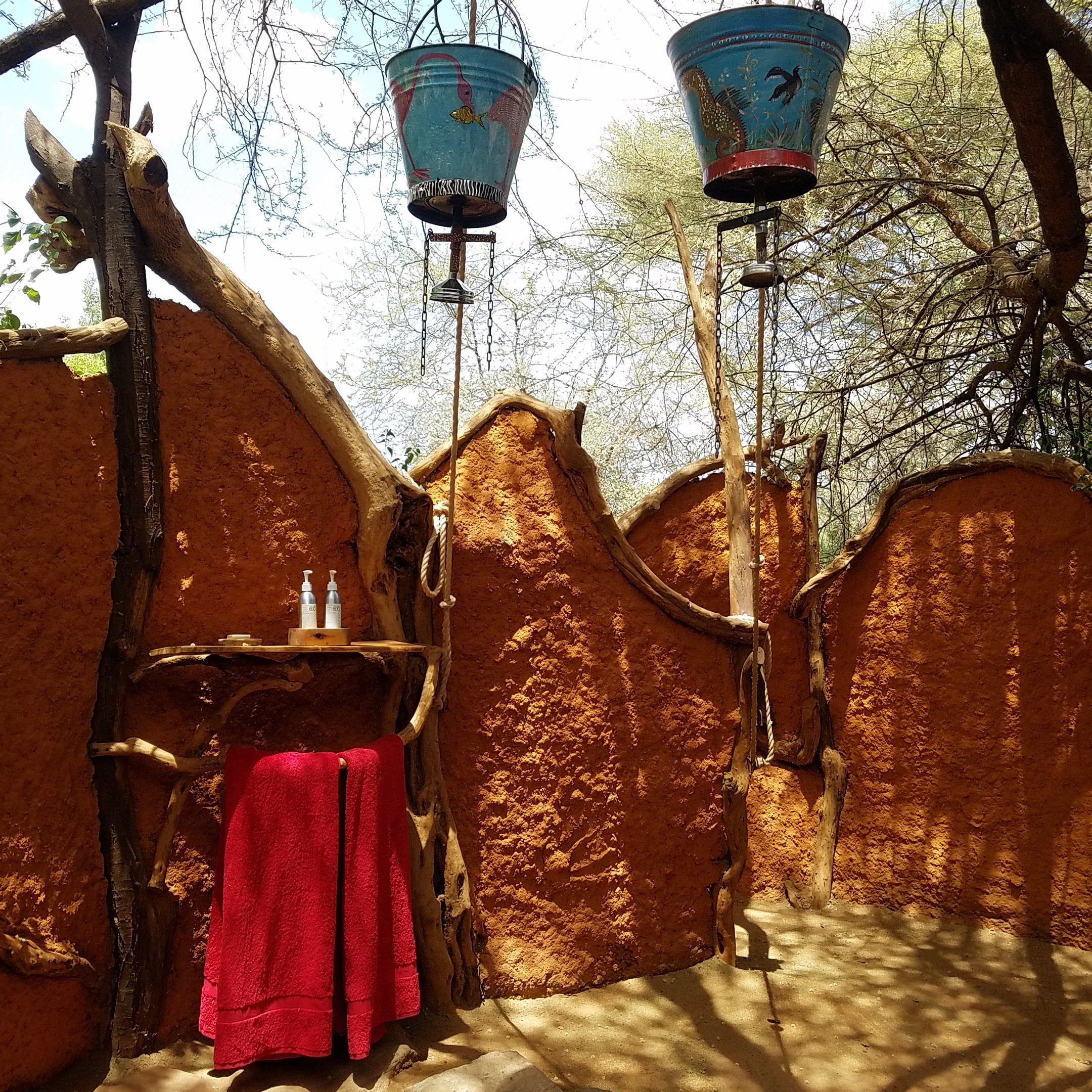 Mud/clay bucket-shower stalls with blue buckets hang overhead, set up outdoors for a camp wash under open sky.