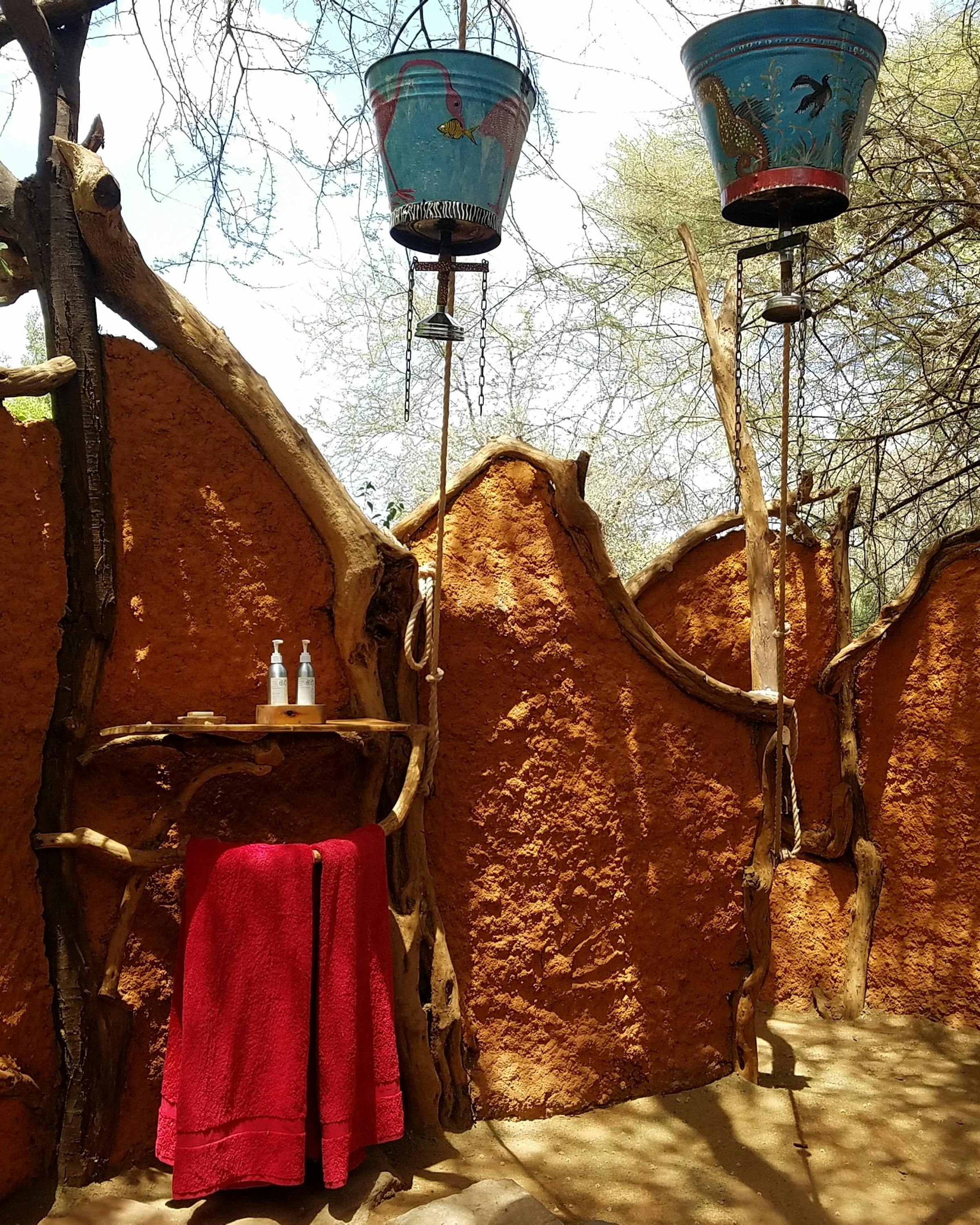 Mud/clay bucket-shower stalls with blue buckets hang overhead, set up outdoors for a camp wash under open sky.