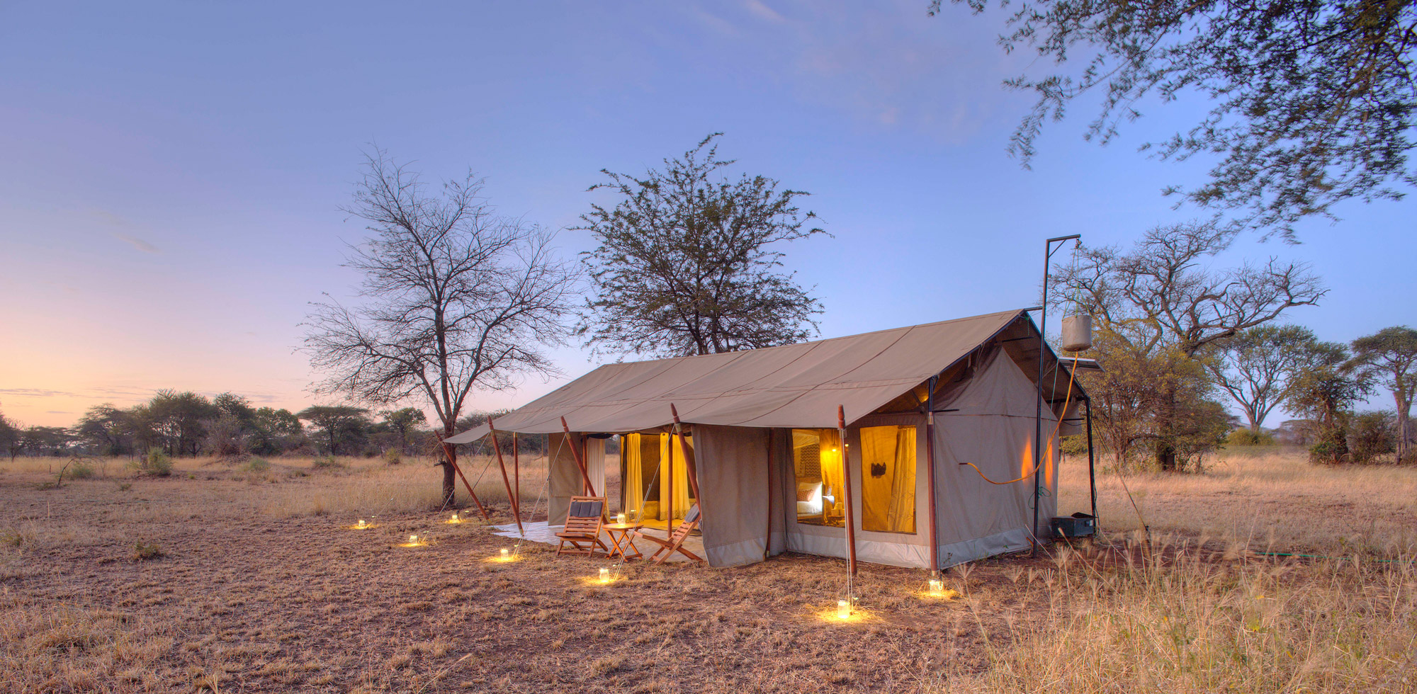Canvas safari guest tent glows at dusk, with a lit porch and nearby trees silhouetted against a pale sky.