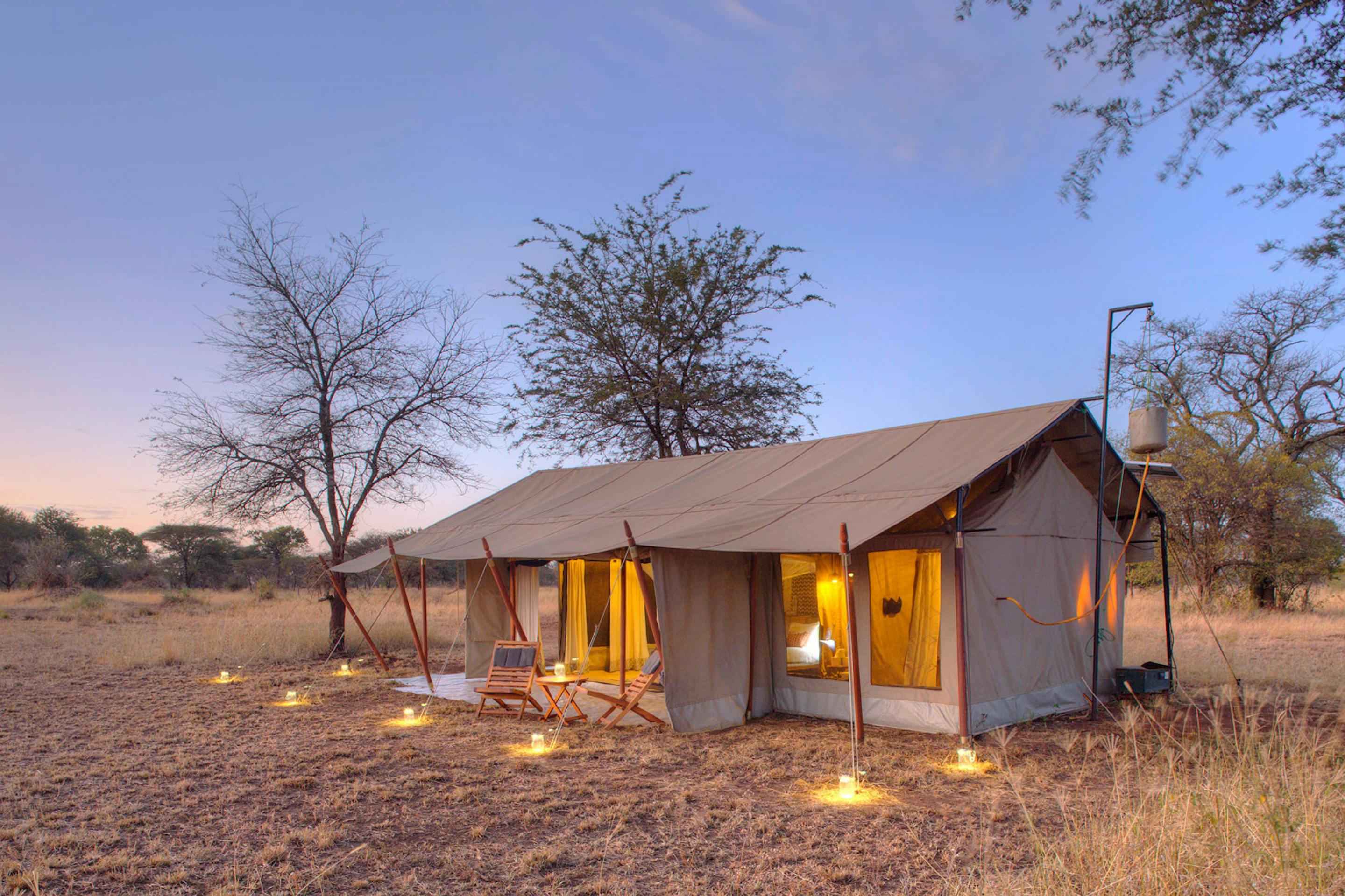 Canvas safari guest tent glows at dusk, with a lit porch and nearby trees silhouetted against a pale sky.