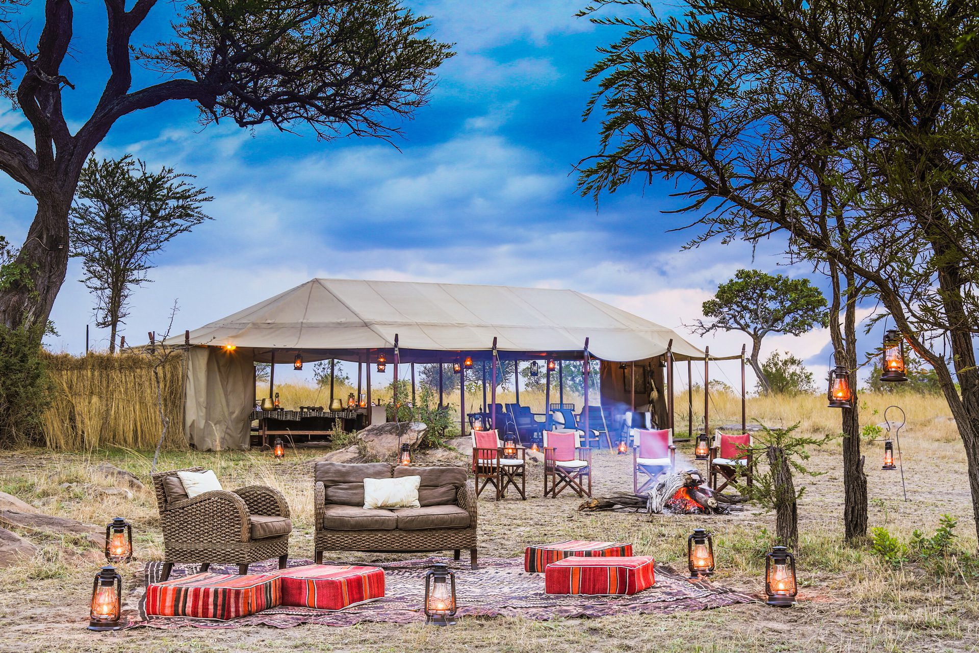 Outdoor safari lounge sits beneath an open tent, with woven chairs, lanterns, rugs, and a bar set on grassland.