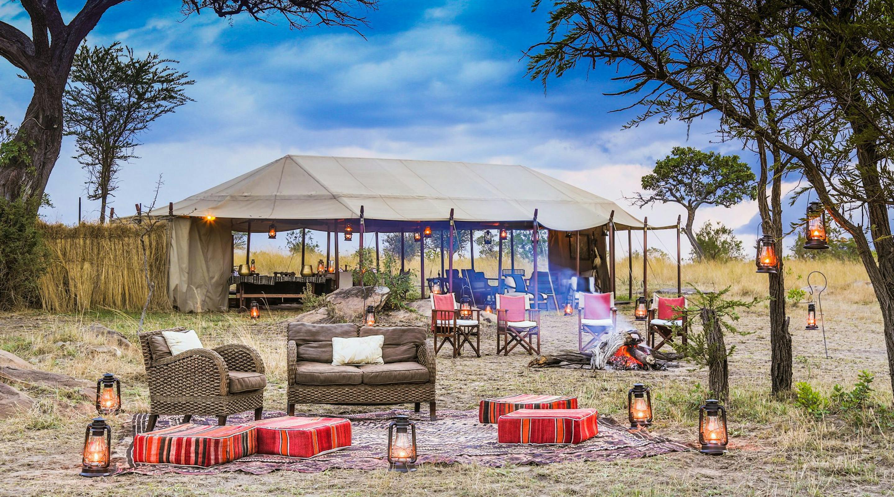 Outdoor safari lounge sits beneath an open tent, with woven chairs, lanterns, rugs, and a bar set on grassland.