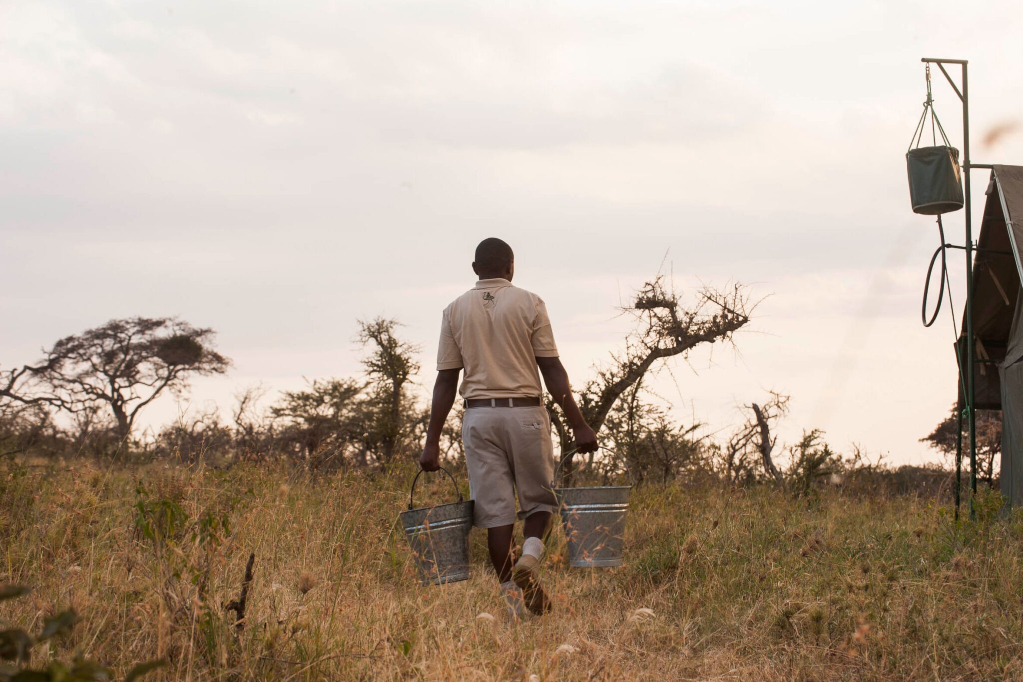 A camp staff member walks across savanna carrying buckets, passing a portable bucket-shower frame in the grass.