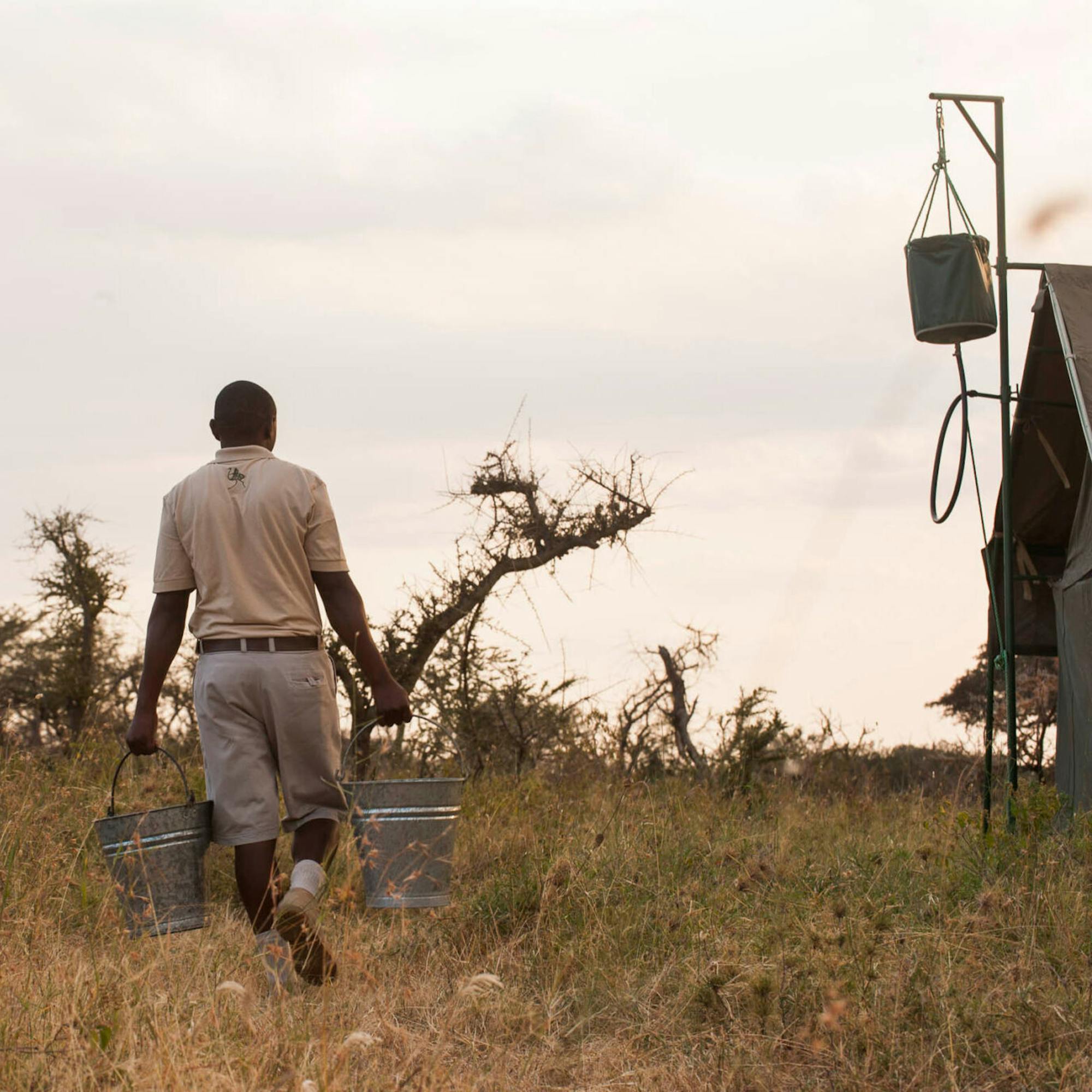 A camp staff member walks across savanna carrying buckets, passing a portable bucket-shower frame in the grass.