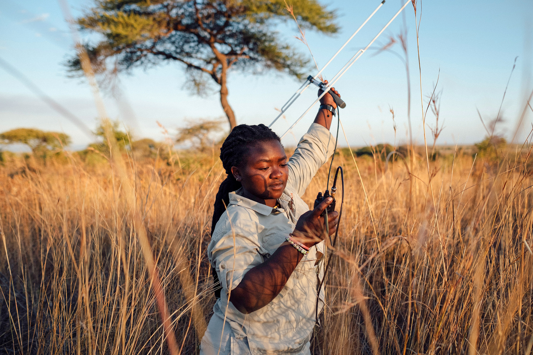 A guide stands in tall golden grass holding a camera, with wide plains and a pale sky stretching behind him.