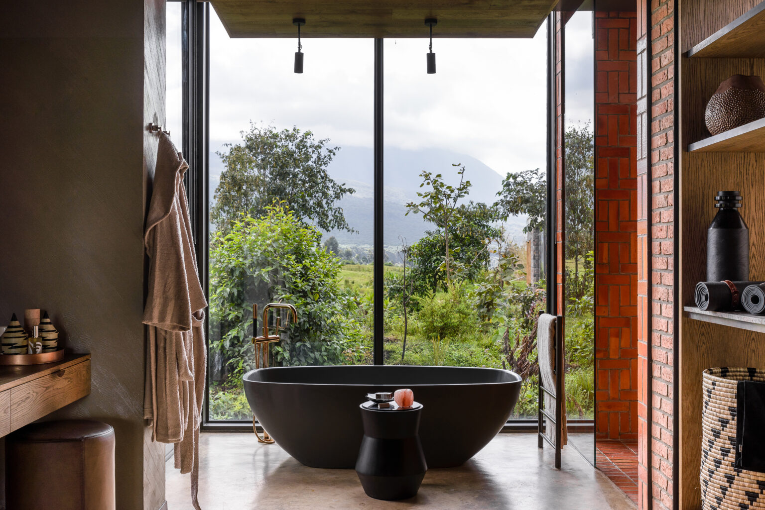 Freestanding black tub sits by a tall window, framed by wood walls and greenery outside under gray skies.