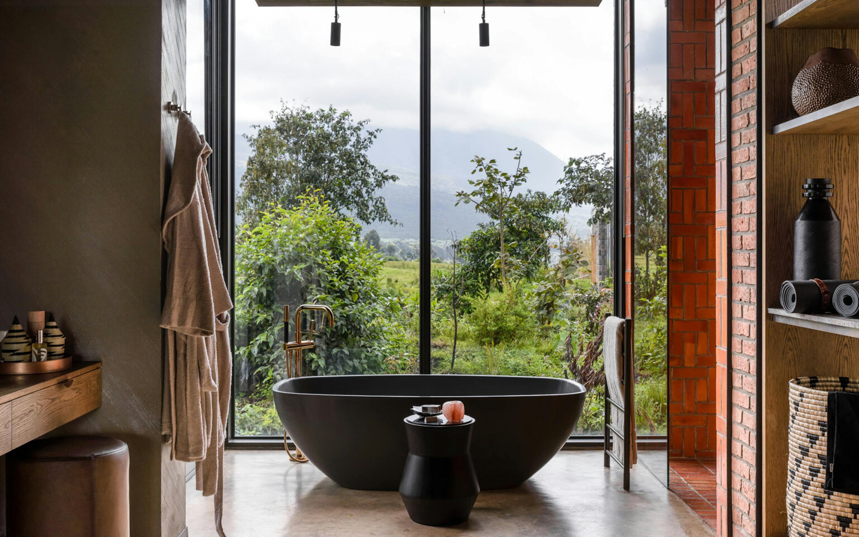 Freestanding black tub sits by a tall window, framed by wood walls and greenery outside under gray skies.