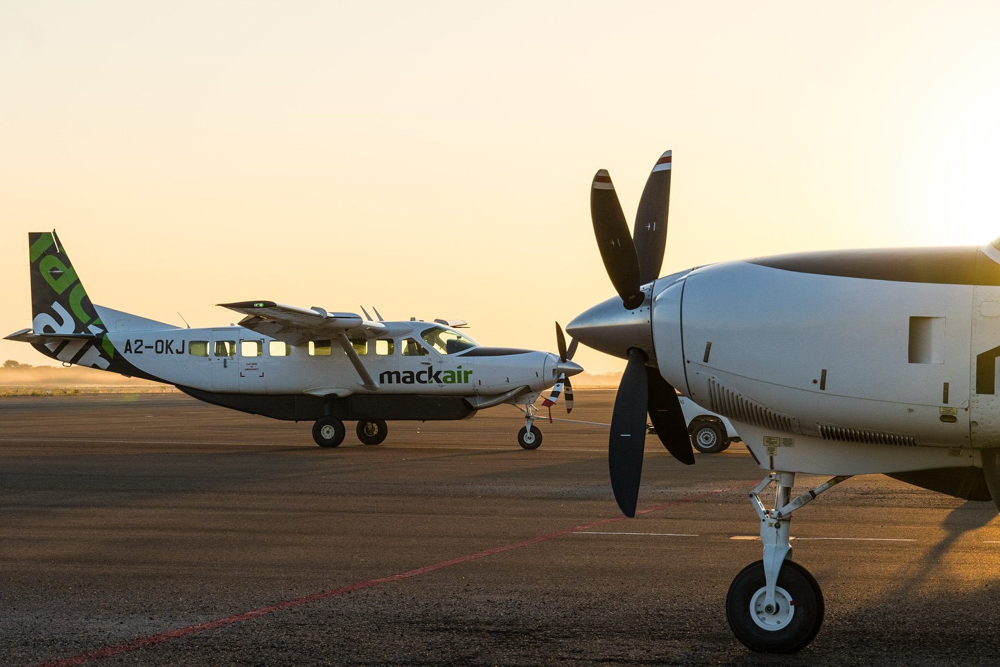 Two small propeller planes sit on a runway at sunset, one wing and spinning propeller filling the foreground.