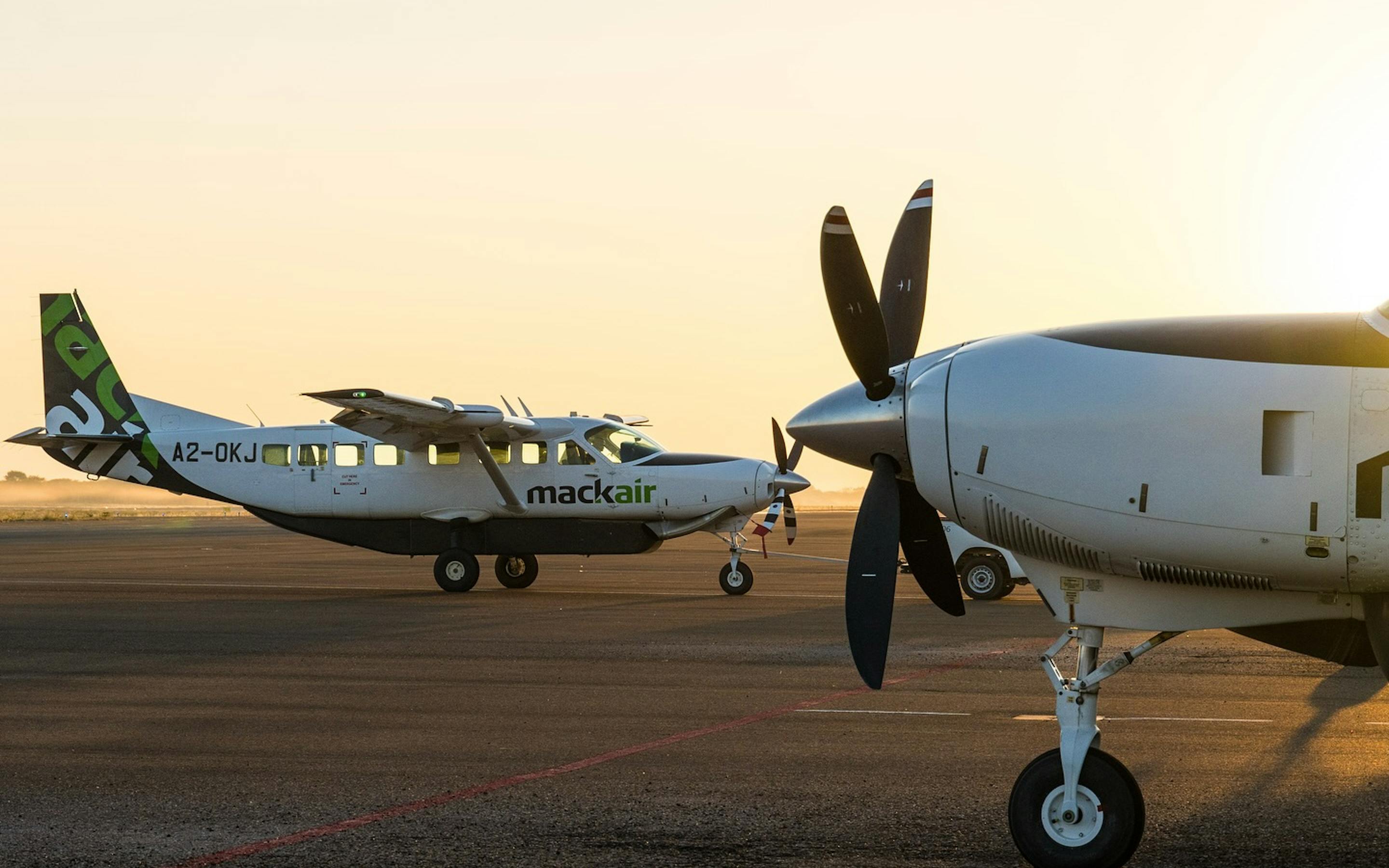 Two small propeller planes sit on a runway at sunset, one wing and spinning propeller filling the foreground.